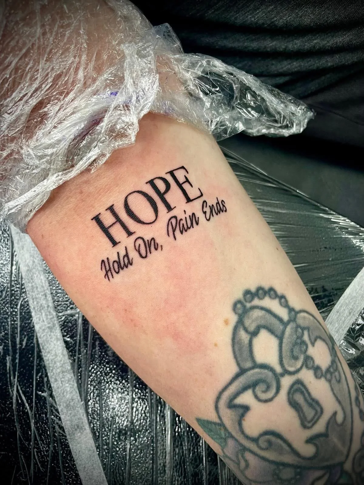Tattoo on person's arm with the word 'HOPE' and the phrase 'Add On, Pain Ends' below it, with a tattoo of a heart with bubble-like accents.