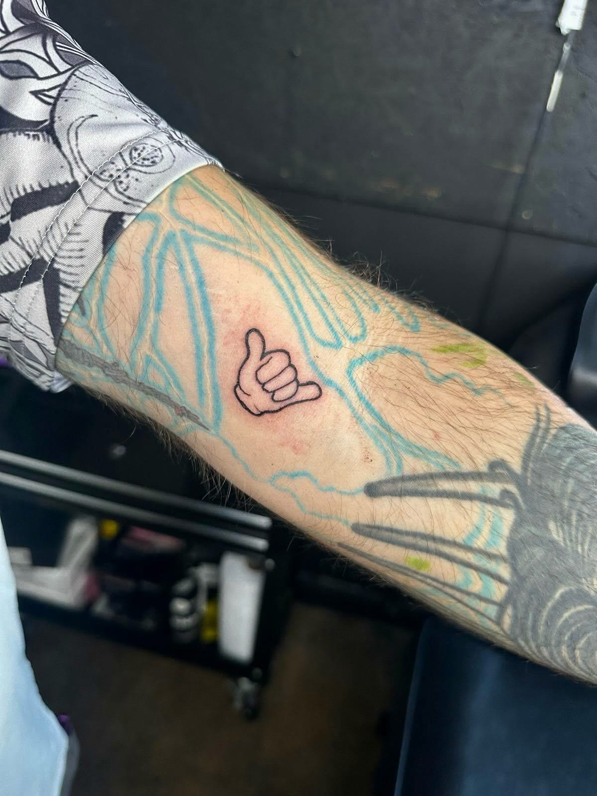 A tattoo on a person's forearm featuring a blue outline of a cartoon hand making a shaka sign, along with other line work. The person is wearing a short-sleeve shirt with a black and white pattern.