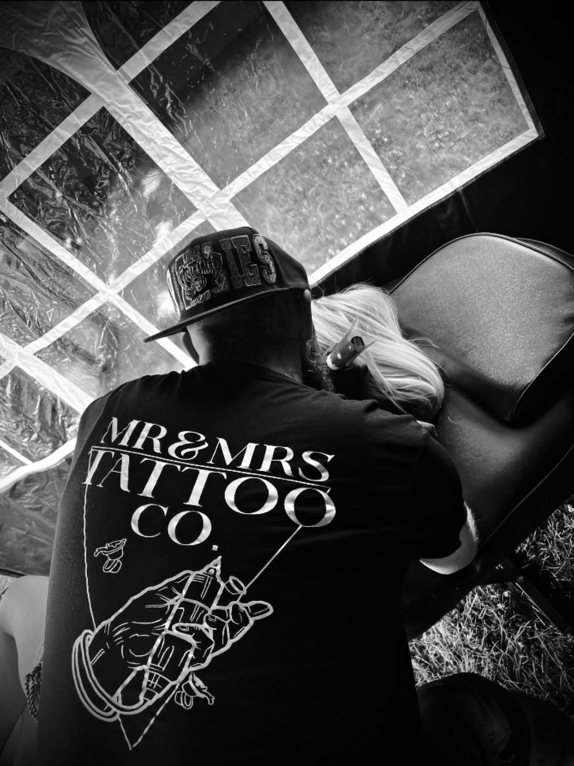A person with a cap and a shirt that says 'MR & MRS TATTOO CO.' is getting a tattoo on a woman's arm or hand, with a work surface and reflective material in the background.