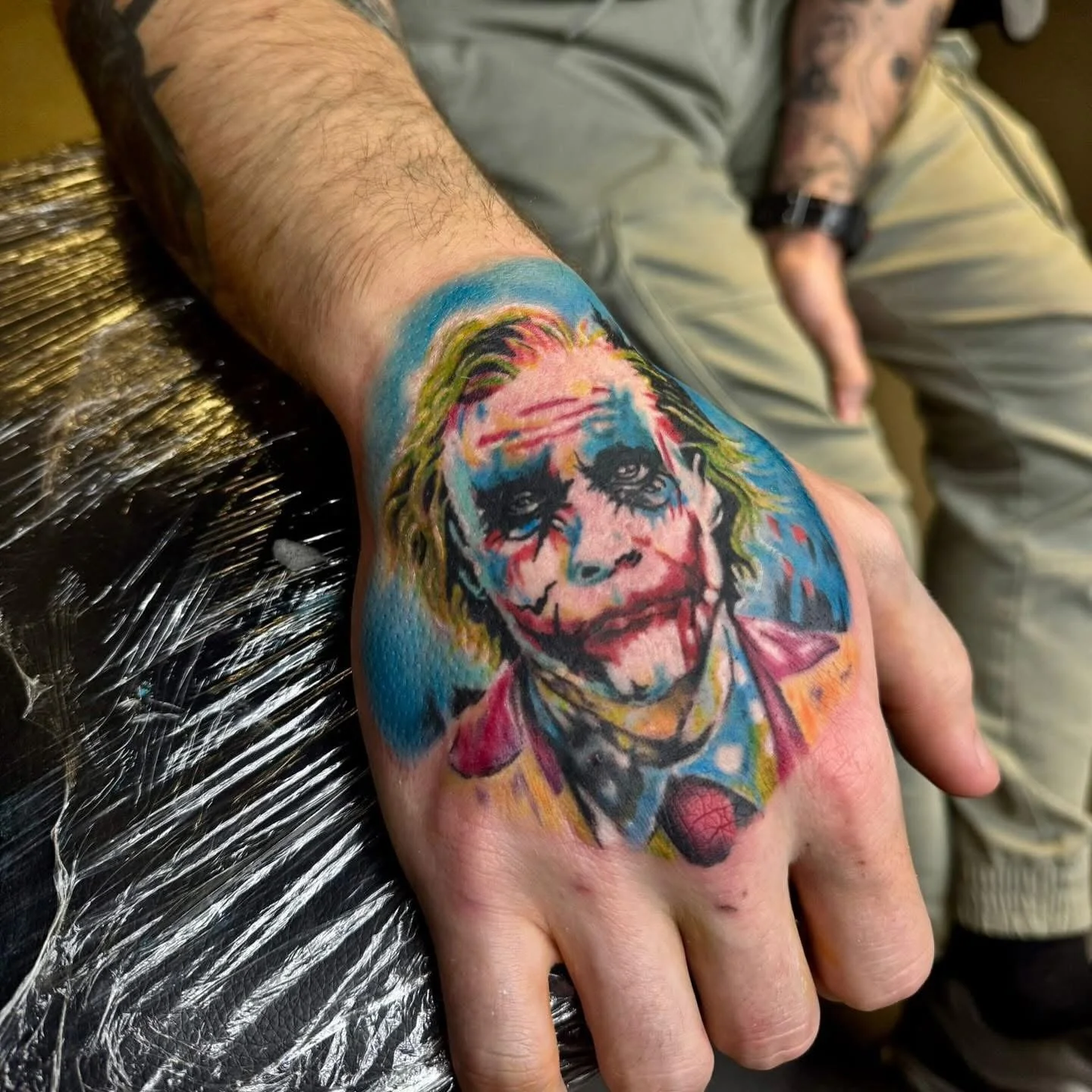 Colorful tattoo of the joker on a person's hand, with a blue background and vibrant colors.