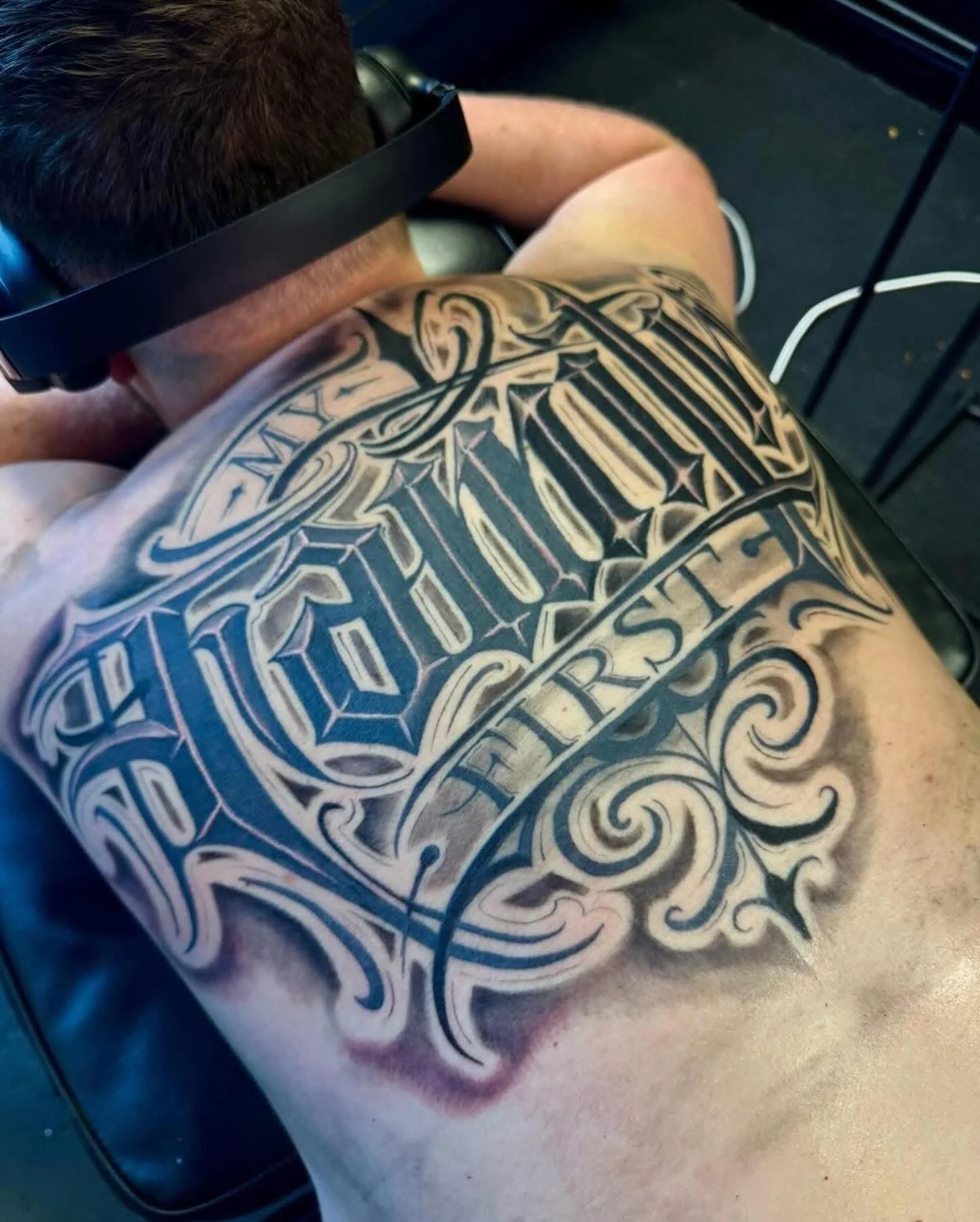 A person's back featuring a large tattoo with intricate black and white lettering and decorative swirling design.