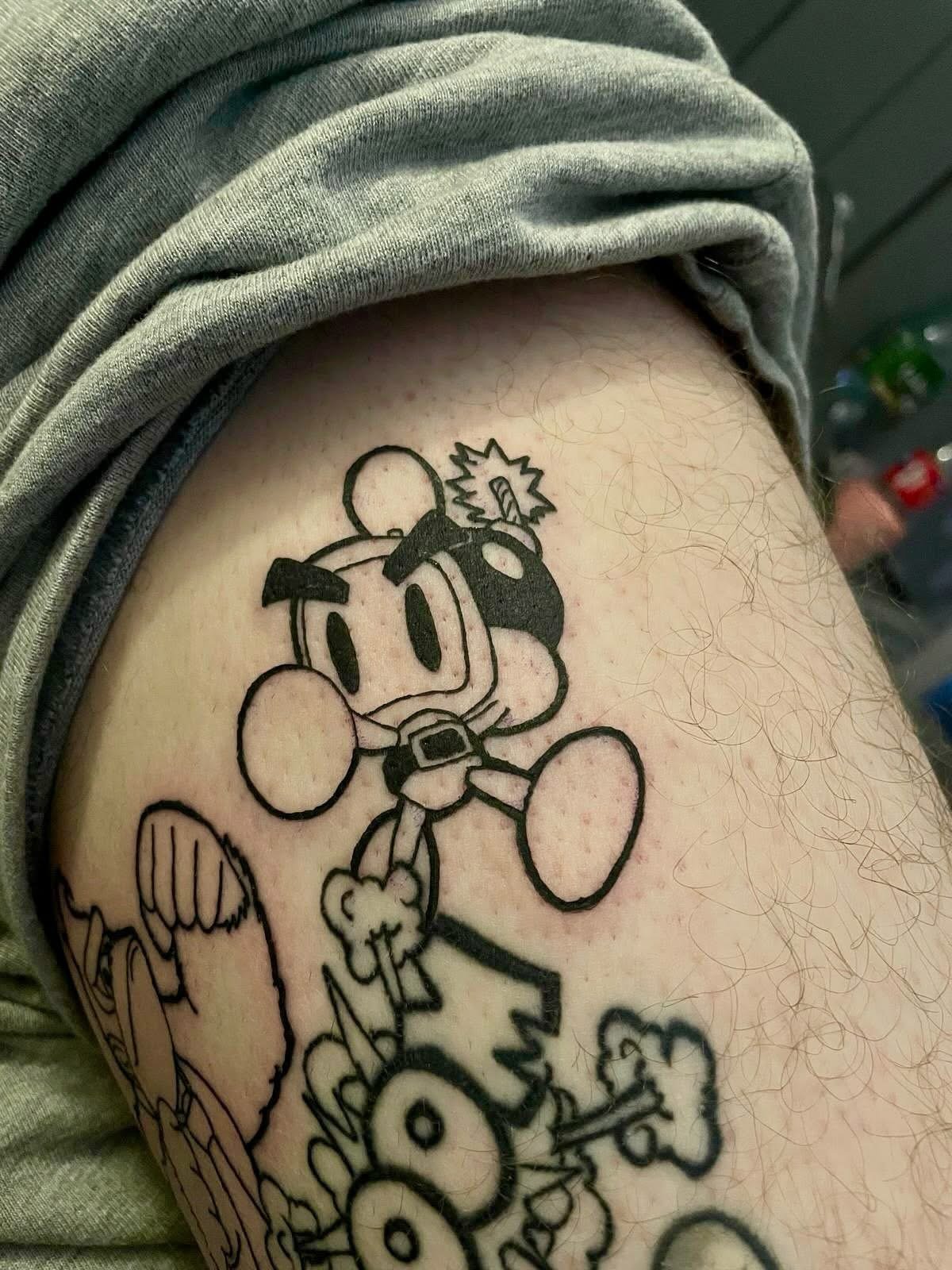 Black line tattoo of Mickey Mouse as a cartoon character with a bomb on his head, accompanied by other cartoon elements, on a person's upper arm.