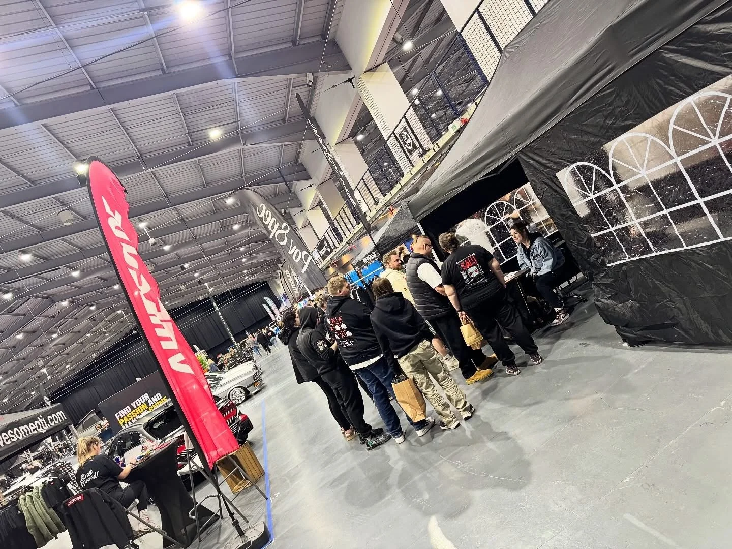 People standing in line inside a large indoor car show or event space, with several booths and displays, including one with a sign reading 'For Service', and a red banner with the word 'WORX'.