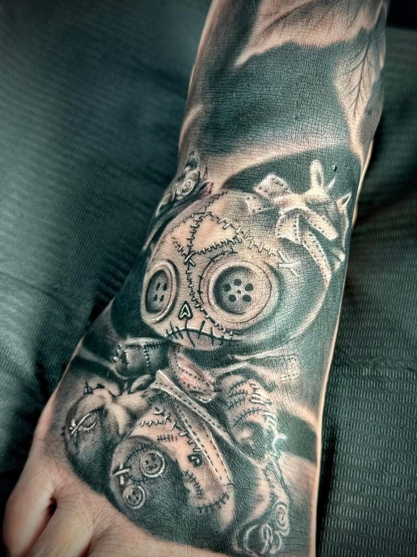A black and gray foot tattoo featuring a stitched teddy bear with button eyes, a zipper mouth, and patchwork details.