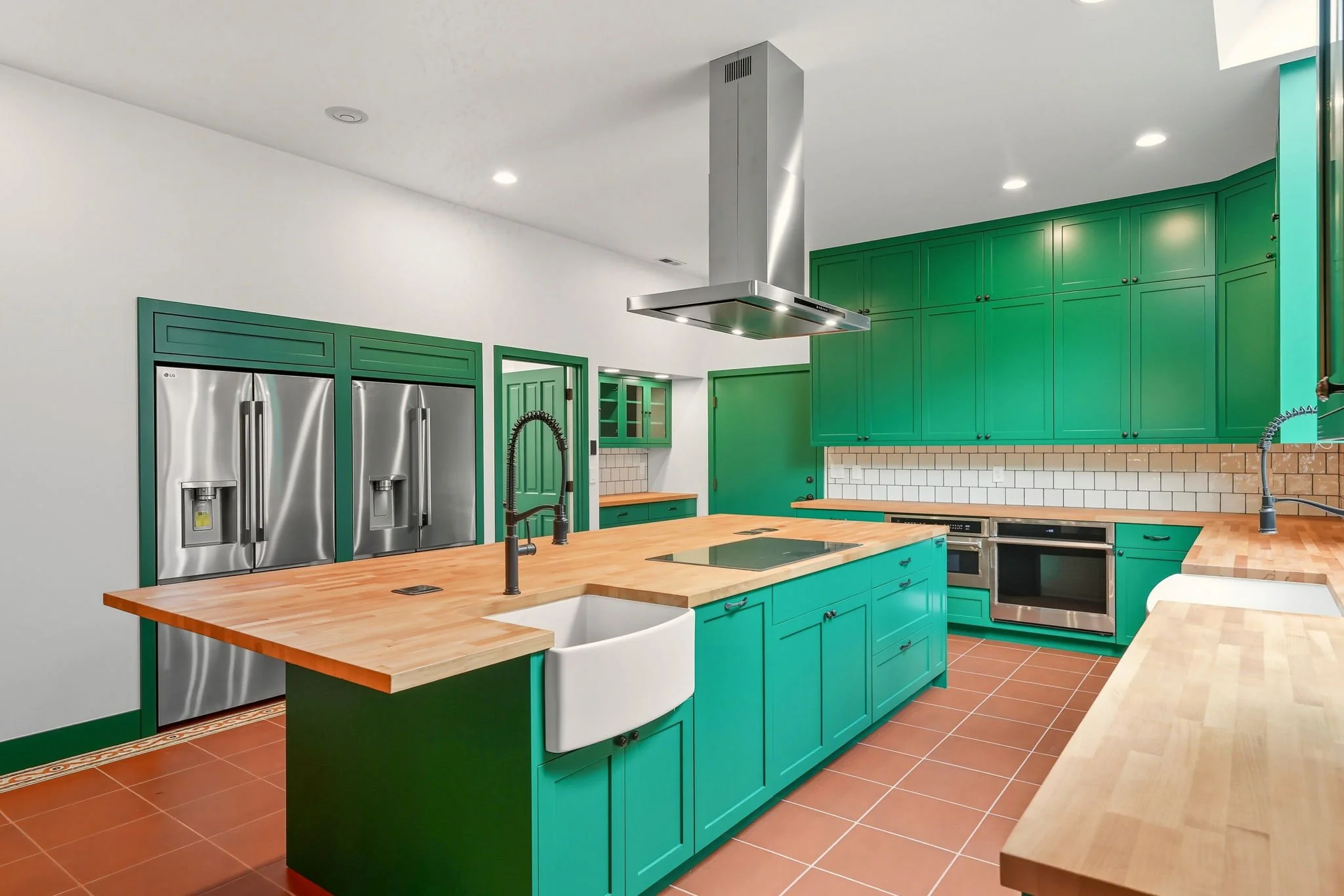 Modern kitchen with green cabinetry, a large wooden island, stainless steel appliances including a double-door refrigerator and oven, and a white farmhouse sink with black fixtures. The kitchen has tiled backsplash and terracotta floor tiles.