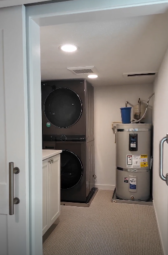 Laundry room remodel with stacked washer and dryer, water heater, cabinet, and laundry supplies. Featuring a custom built sliding white barn door.