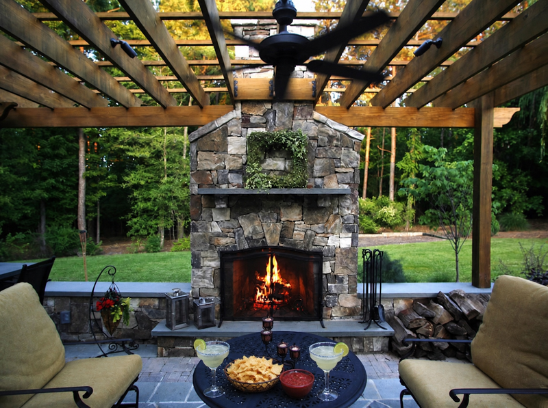 Outdoor patio with a stone fireplace, fire burning inside, and a pergola overhead. Two chairs with cushions face the fireplace. A small table holds snacks, drinks, and candles. Trees and a lawn are visible in the background.