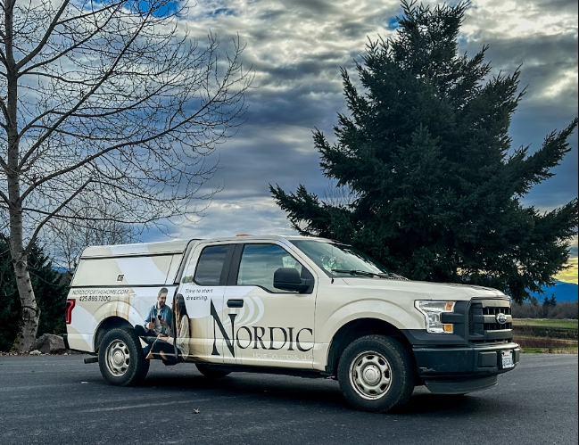 White pickup truck with ads for Nordic travel company, parked on the road, with leafless trees and a large evergreen tree under a cloudy sky in the background.