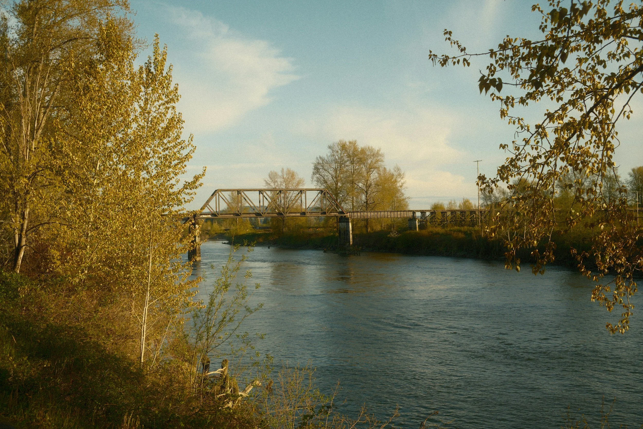 A scenic view of the river in downtown Snohomish, WA with trees on both sides and a bridge crossing over it, under a partly cloudy sky.
