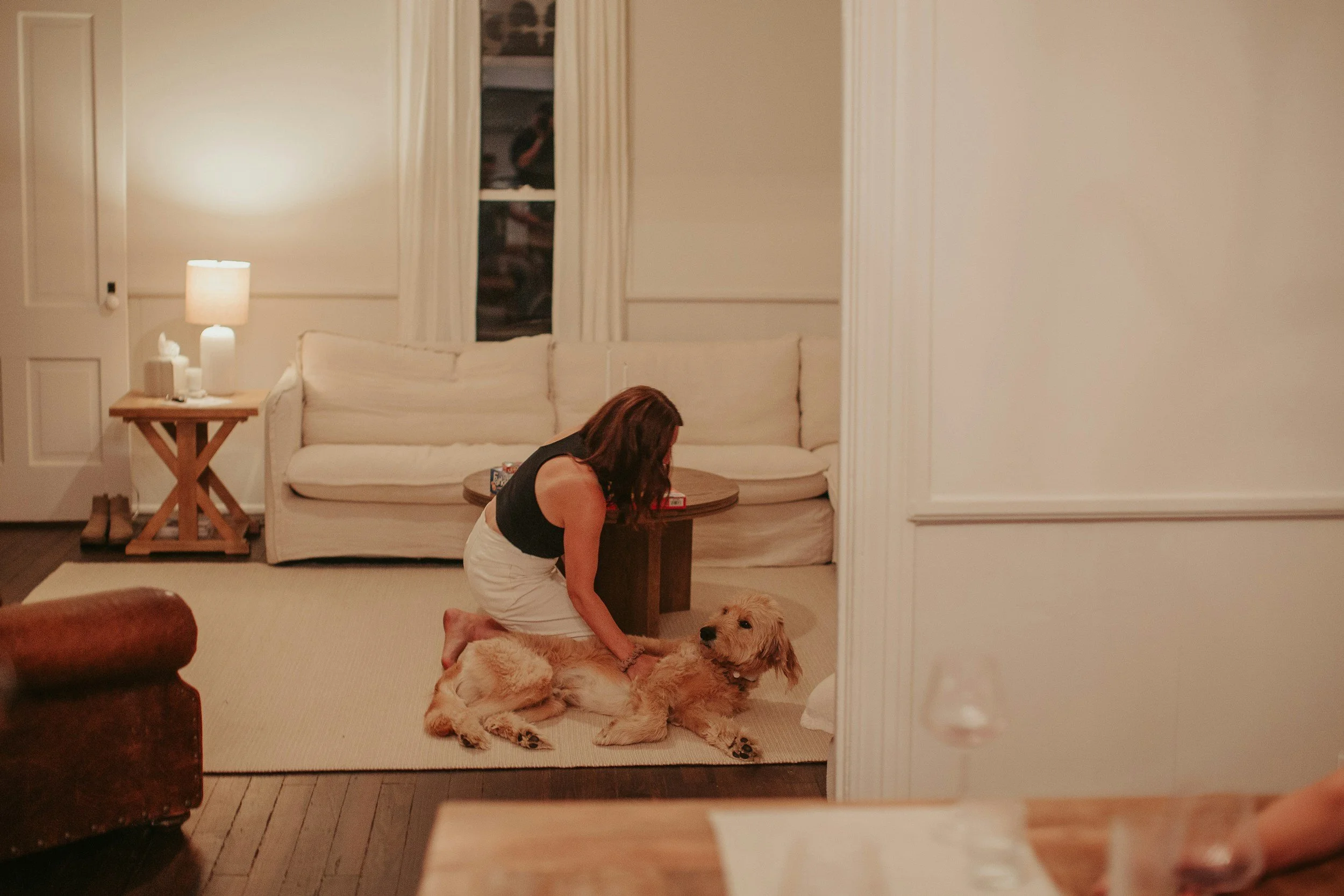 A woman at home in hew newly remodeled dining room kneeling on a beige carpet, playing with a large golden retriever dog lying on the floor.