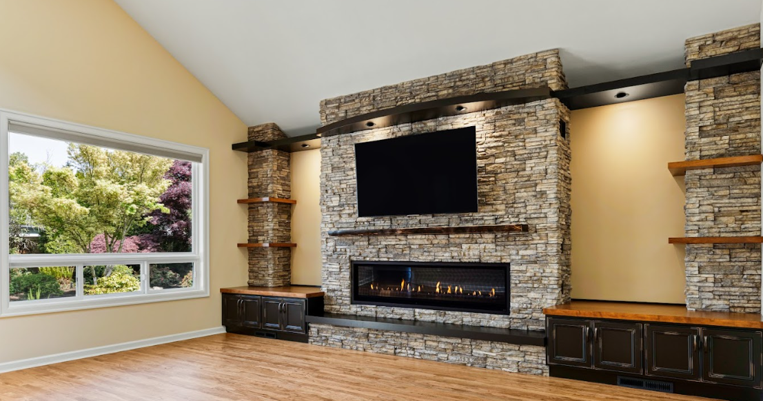 Living room remodel with vaulted ceilings and a stone fireplace, flat-screen TV above a wooden mantel, black built-in cabinets on either side, and built-in shelves on stone pillars, with hardwood floors and windows on either side.