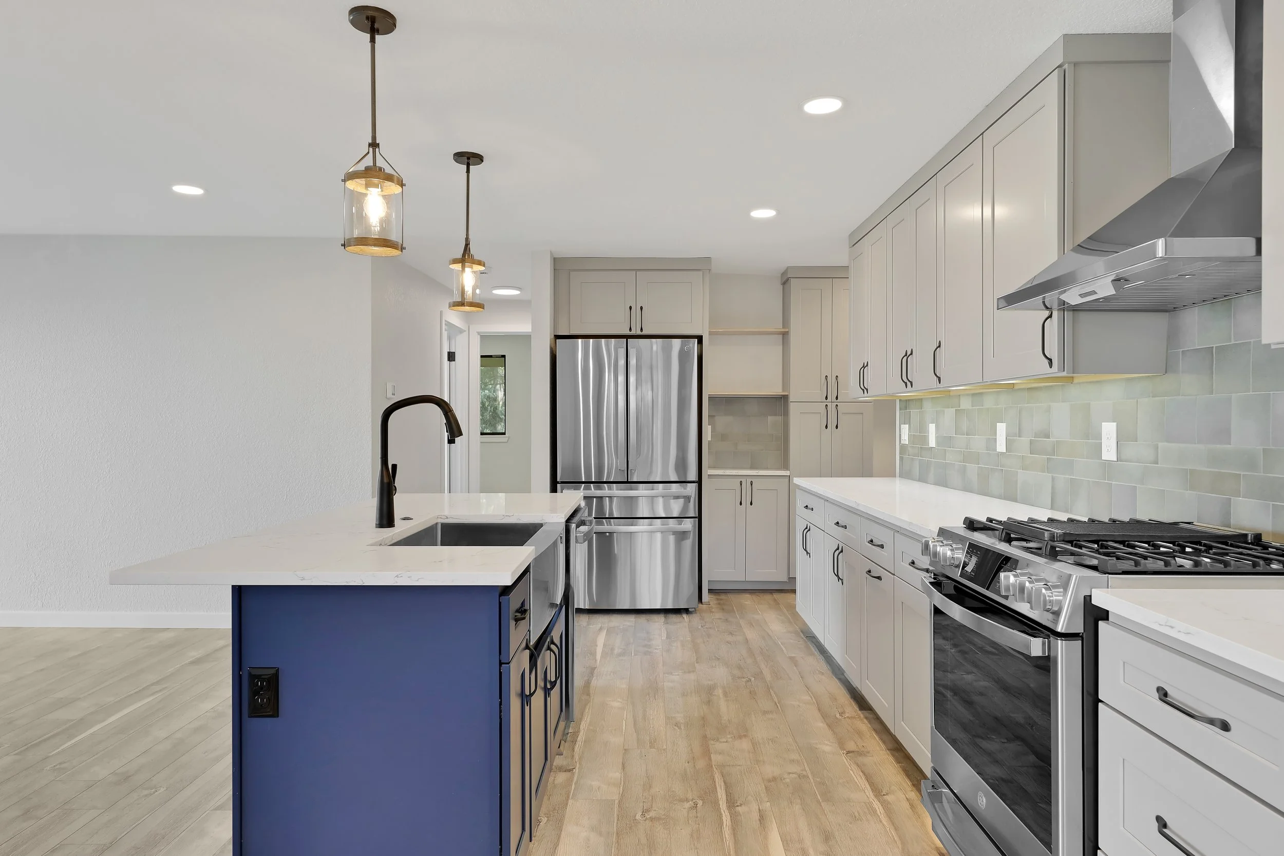 Side view of a Modern kitchen with light gray cabinets, stainless steel refrigerator, and a blue island with a black faucet, pendant lights 