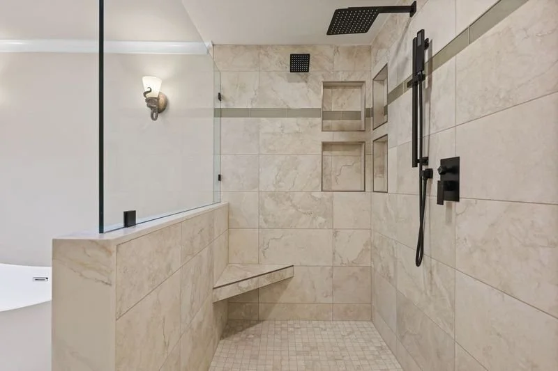 A custom built walk-in shower with beige marble tiles, two black rain showerheads, a handheld shower, built-in wall niches, a glass divider, and a built-in bench. Design-build completed by Nordic Home Solutions in Monroe, WA.
