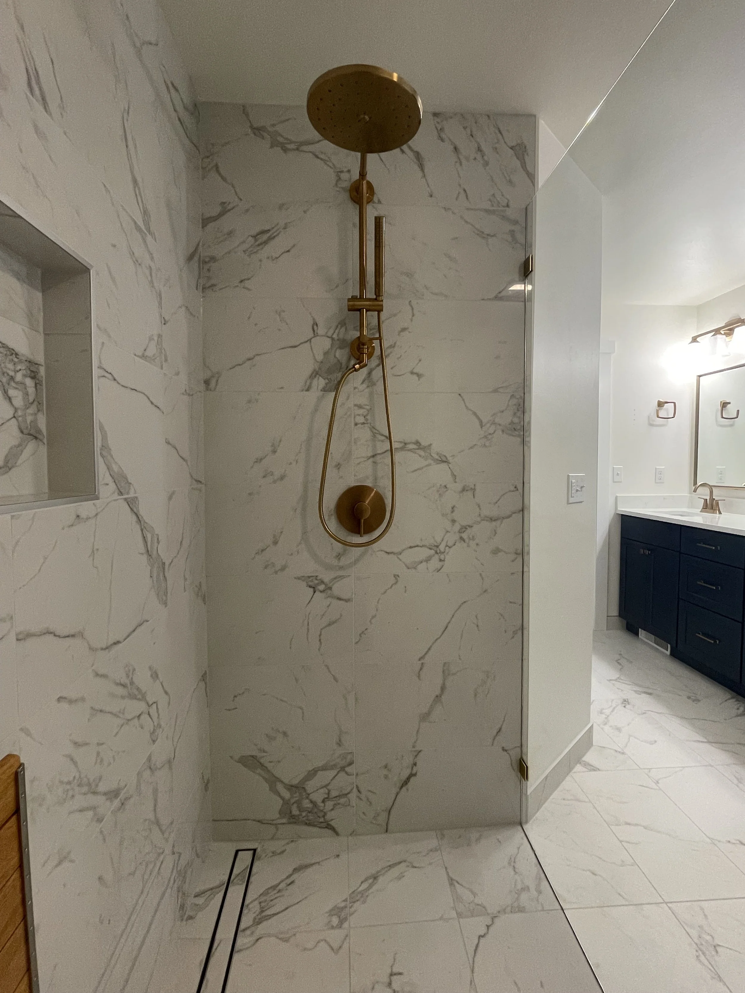 Custom walk-in shower in a Monroe, WA primary bathroom remodel featuring floor-to-ceiling marble-look tile, a rainfall showerhead, handheld shower and brushed gold fixtures.