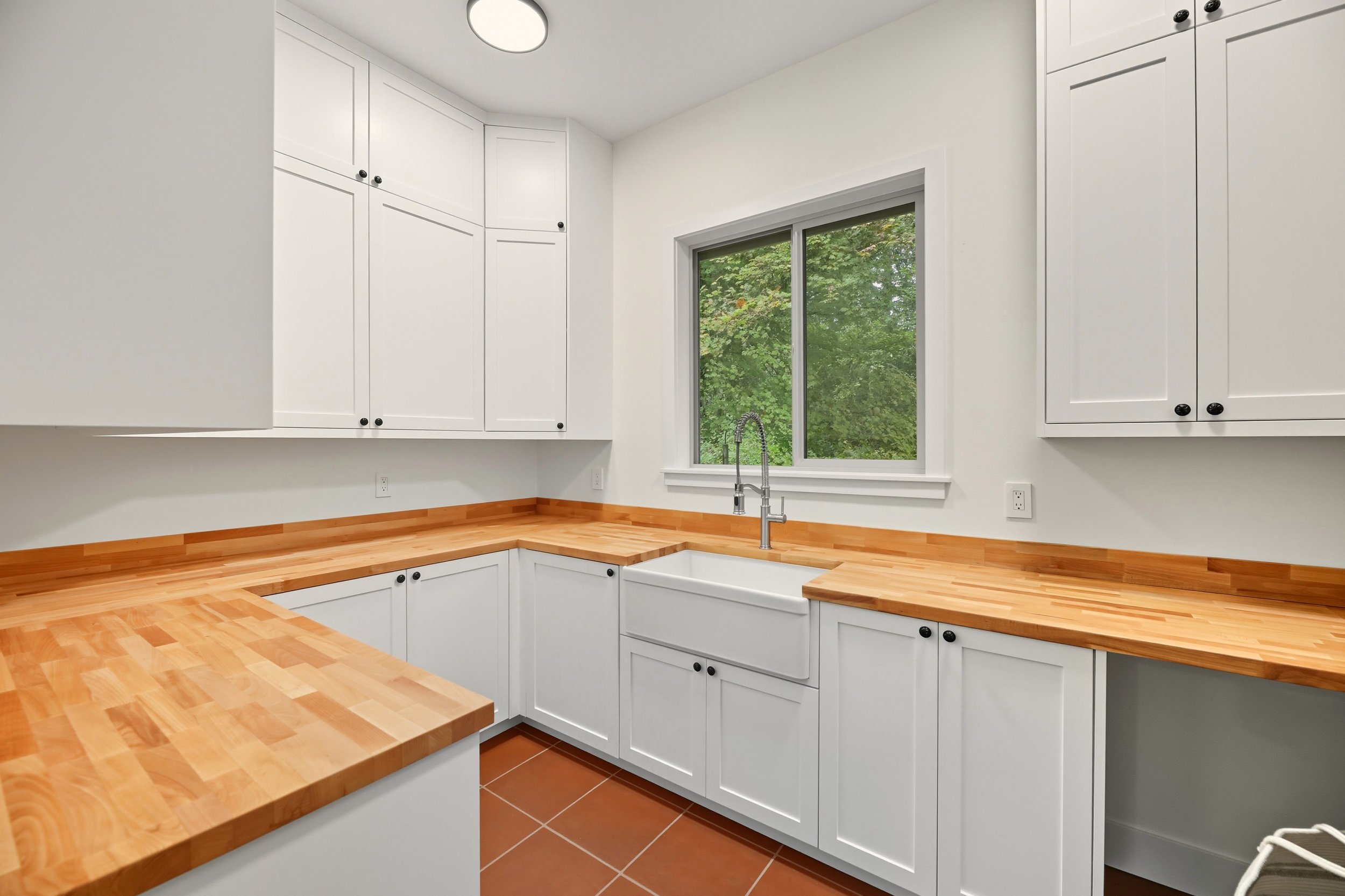 Custom built laundry room with white storage cabinets, a window above a white farmhouse sink, butcher block  countertops, terracotta tile flooring