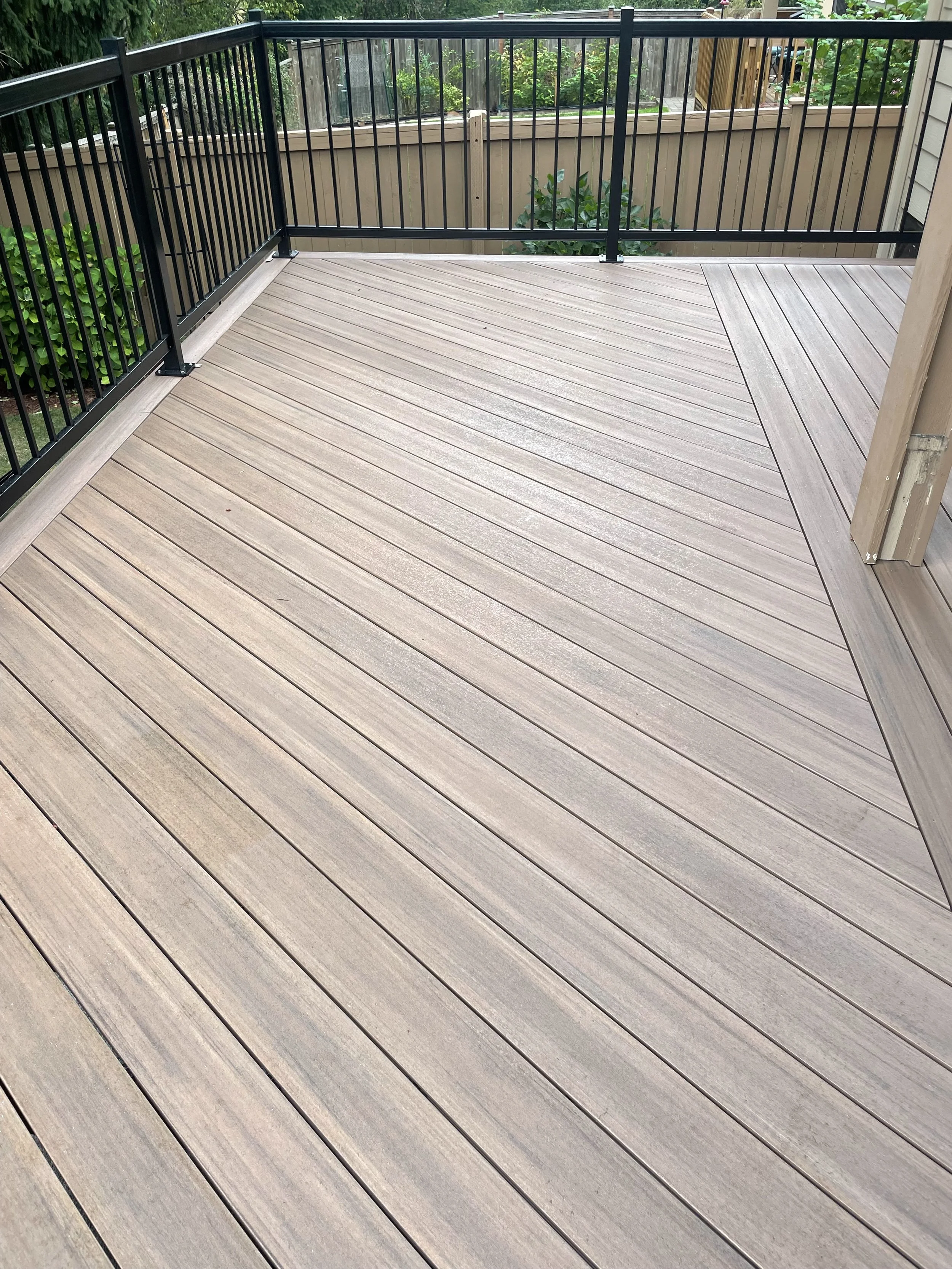A wooden deck with a black metal railing surrounding it, shown from an outdoor area during daylight. Some greenery is visible beyond the railing.