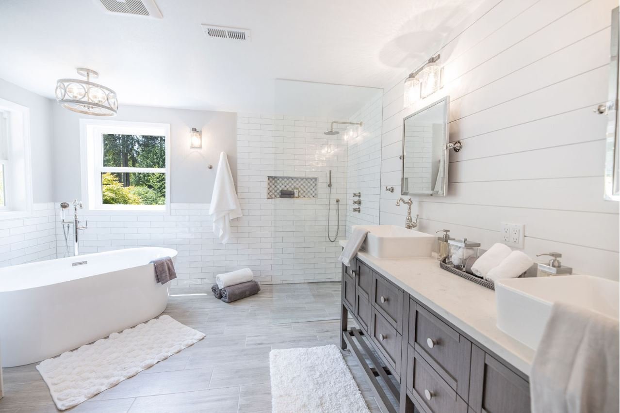Bright white newly remodeled bathroom designed by Nordic Home Solutions. Featuring a freestanding  bathtub, double sinks, and a walk-in shower with a glass partition. There are towels, a bath mat, and a window overlooking backyard greenery.