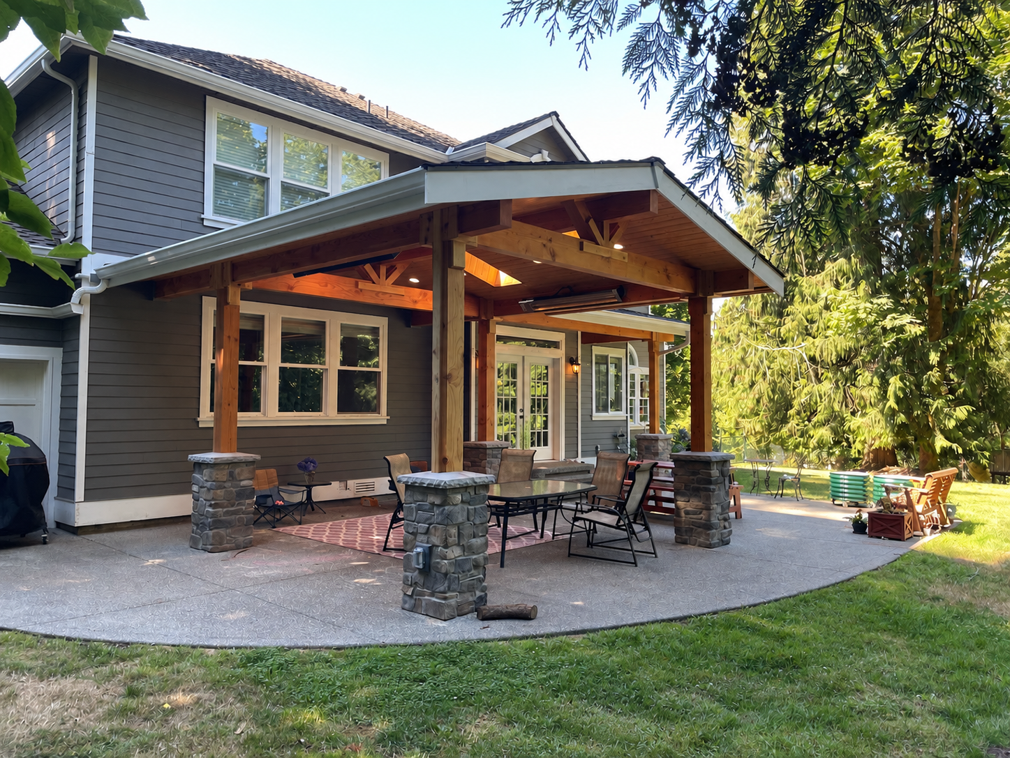 Backyard patio with a covered seating area, outdoor table and chairs, surrounded by greenery and trees, with the house in the background.