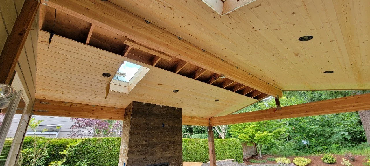 Interior view of a house under construction with unfinished wooden ceiling and roof, skylight, and surrounding greenery visible outside.
