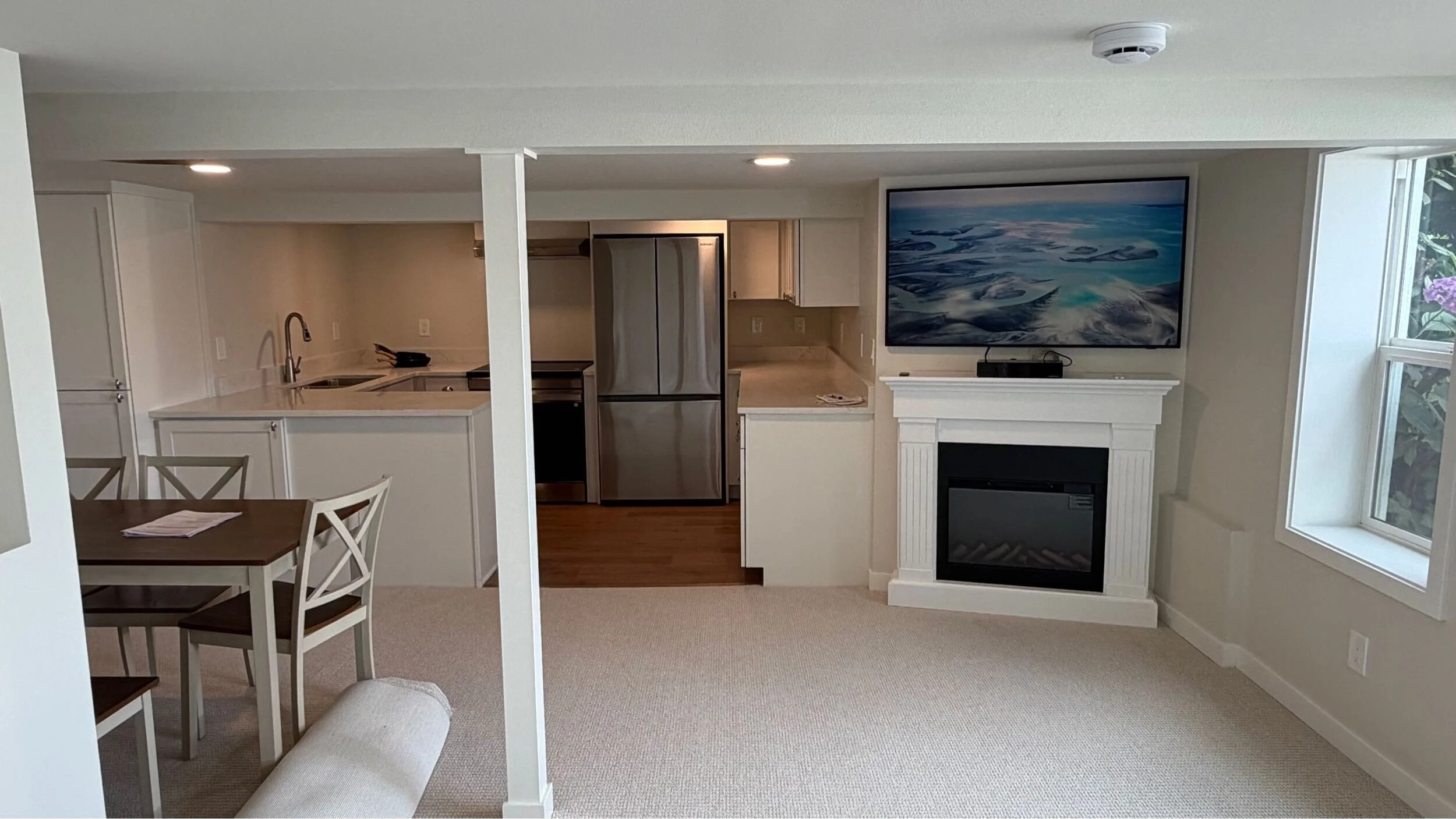A fully remodeled downstairs bonus suite showcasing an open concept living space with an electric fireplace, mounted tv, and modern white kitchen.