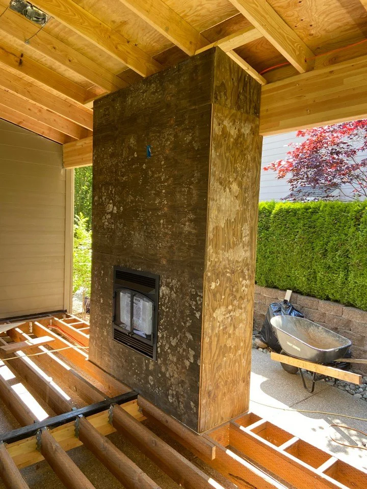 Construction site featuring a large vertical wooden fireplace with a mounted insert, surrounded by unfinished wooden flooring and ceiling. A wheelbarrow and construction tools are visible outside.