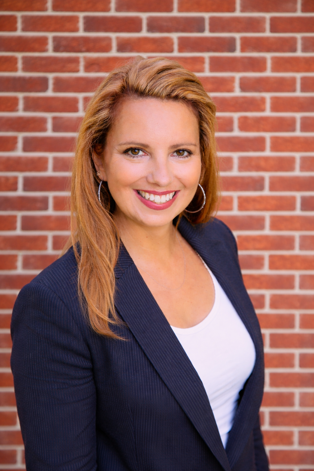 A woman with reddish-brown hair smiling, wearing a navy blazer and a white top, standing in front of a brick wall.