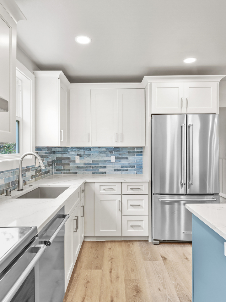 Modern kitchen remodel showcasing white kitchen cabinets, with marble countertops, blue tile backsplash, stainless steel appliances, and a window.
