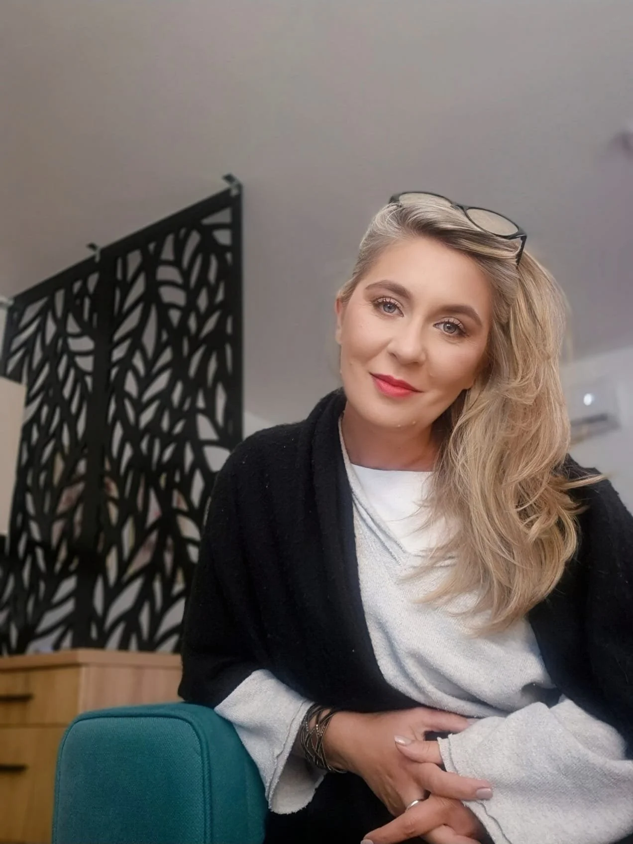 A woman with blonde hair, glasses on her head, sitting at a table, smiling, wearing a white shirt and black cardigan.
