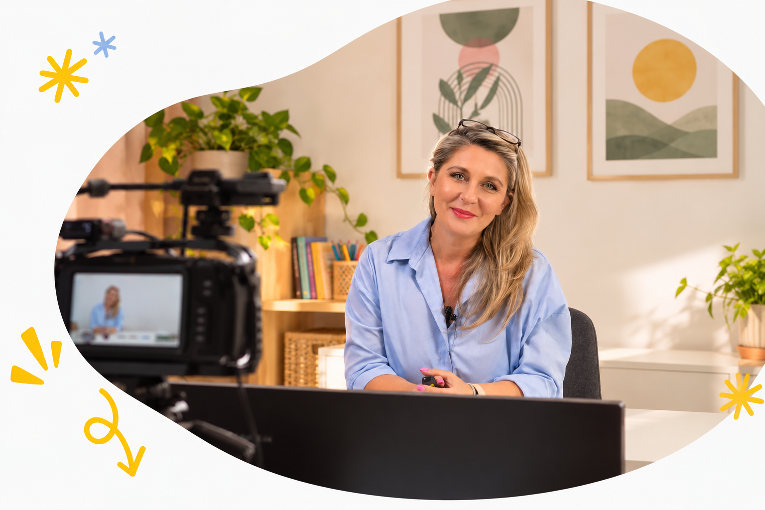 A woman in a blue shirt filming a video in a well-lit room with plants and framed art on the wall.