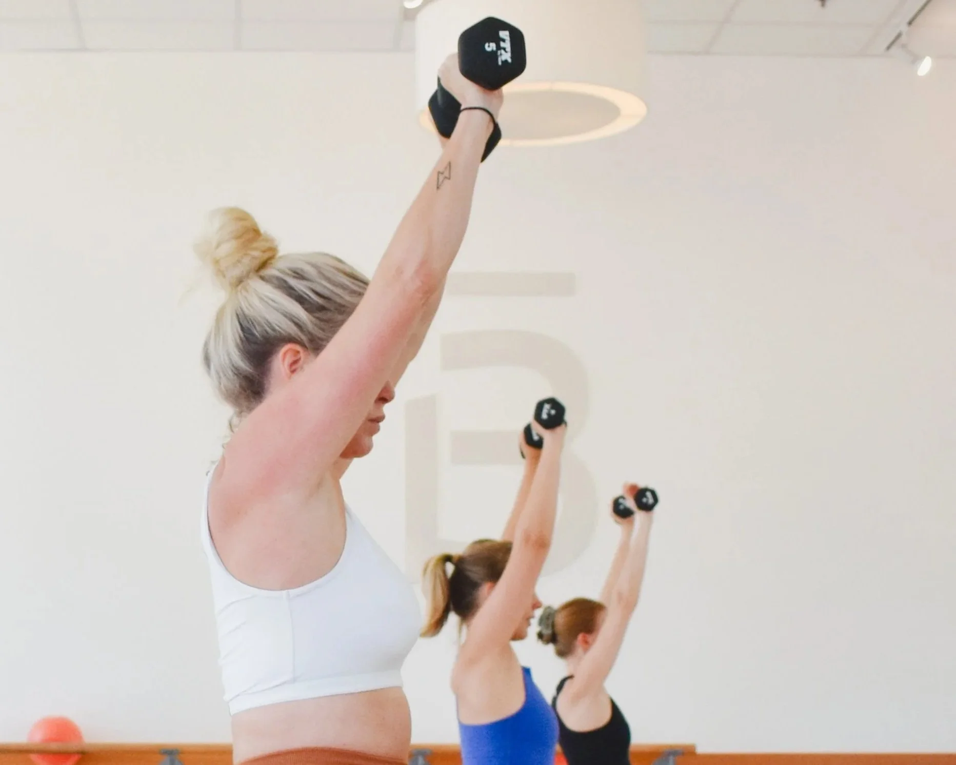 Women participating in a fitness class lifting dumbbells overhead in a gym or studio.