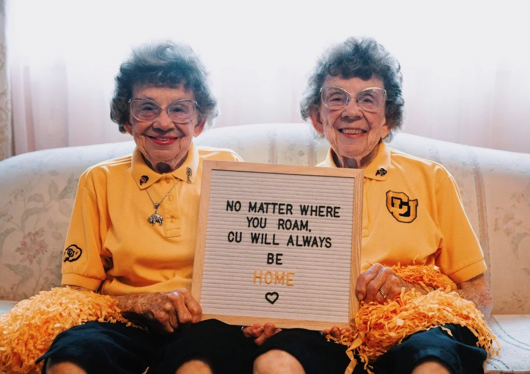 Two elderly women with glasses, wearing yellow CU shirts, smiling while sitting on a beige patterned couch, holding a letter board with the message 'No matter where you roam, CU will always be home' and a small heart.