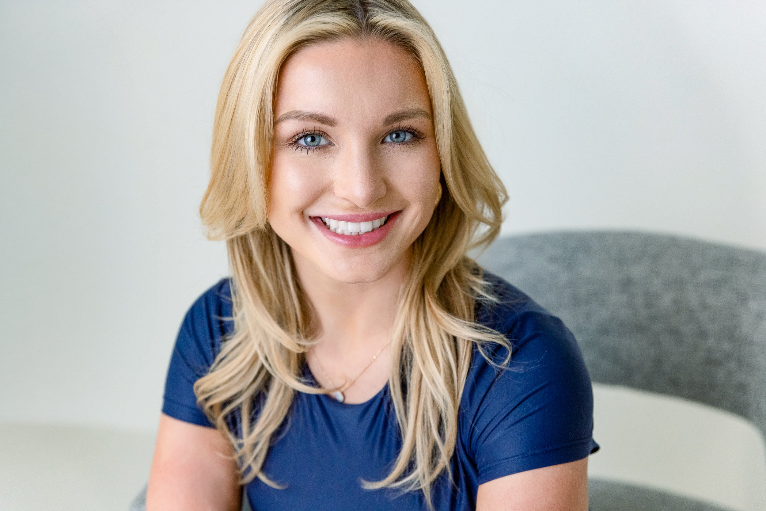Smiling woman in blue top and blonde hair, seated in a gray studio chair during a professional headshot session in Colorado Springs.
