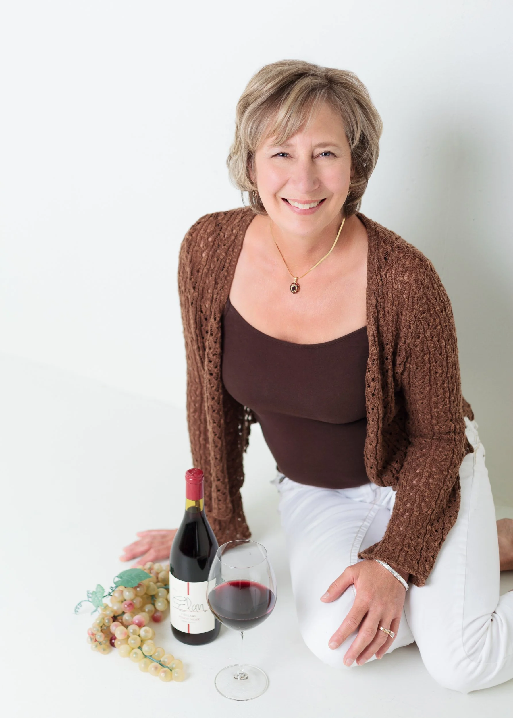 Branding portrait of a smiling woman seated on a white backdrop, styled with wine, grapes, and elegant jewelry. Captured by Colorado Springs photographer Charlotte Harloff, this session blends professionalism, personality and a relaxed lifestyle.