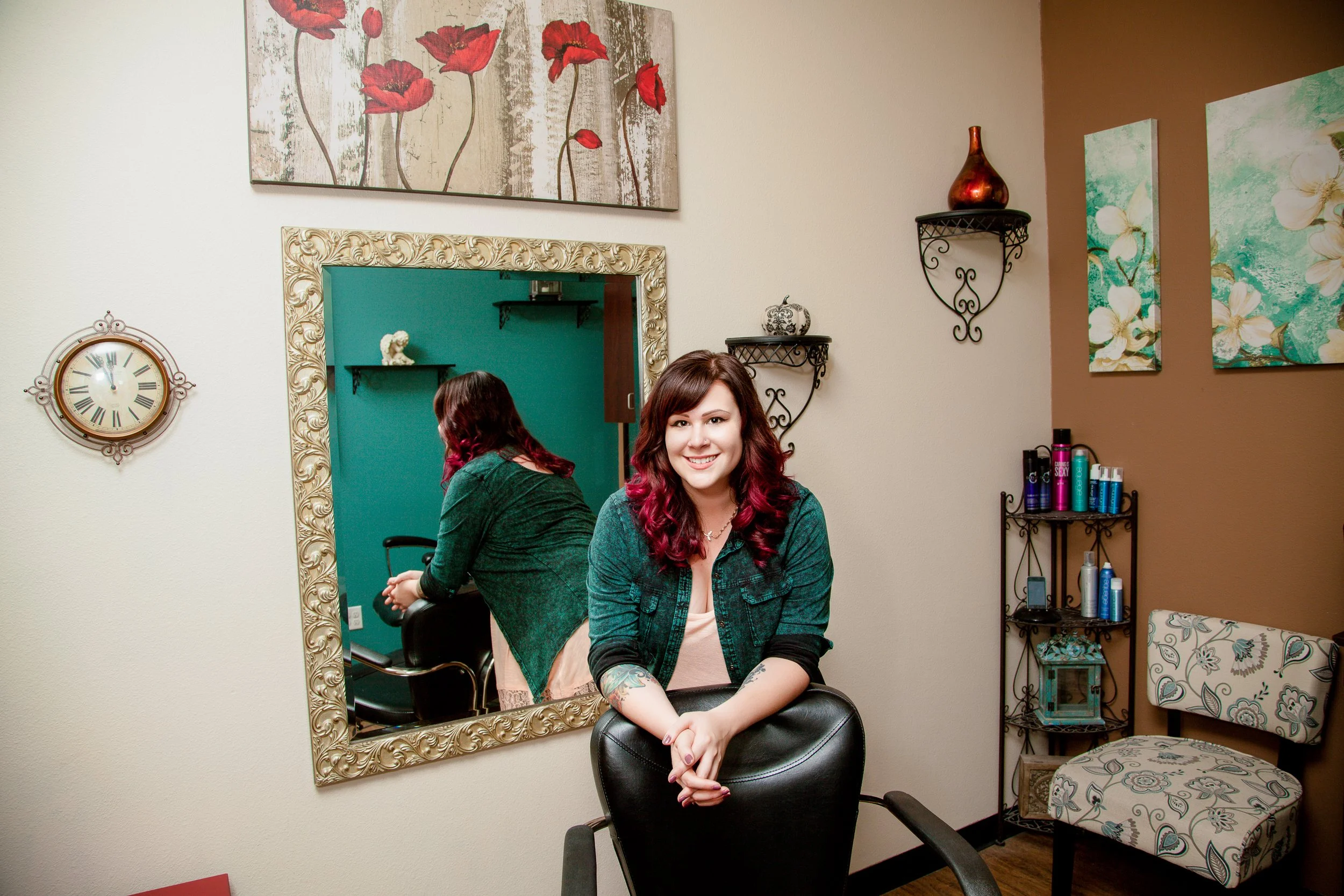Smiling hairdresser with dark brown and pink hair, leaning on a black salon chair in a styled Colorado Springs salon, surrounded by wall art and shelves of hair products during a branding photo shoot.