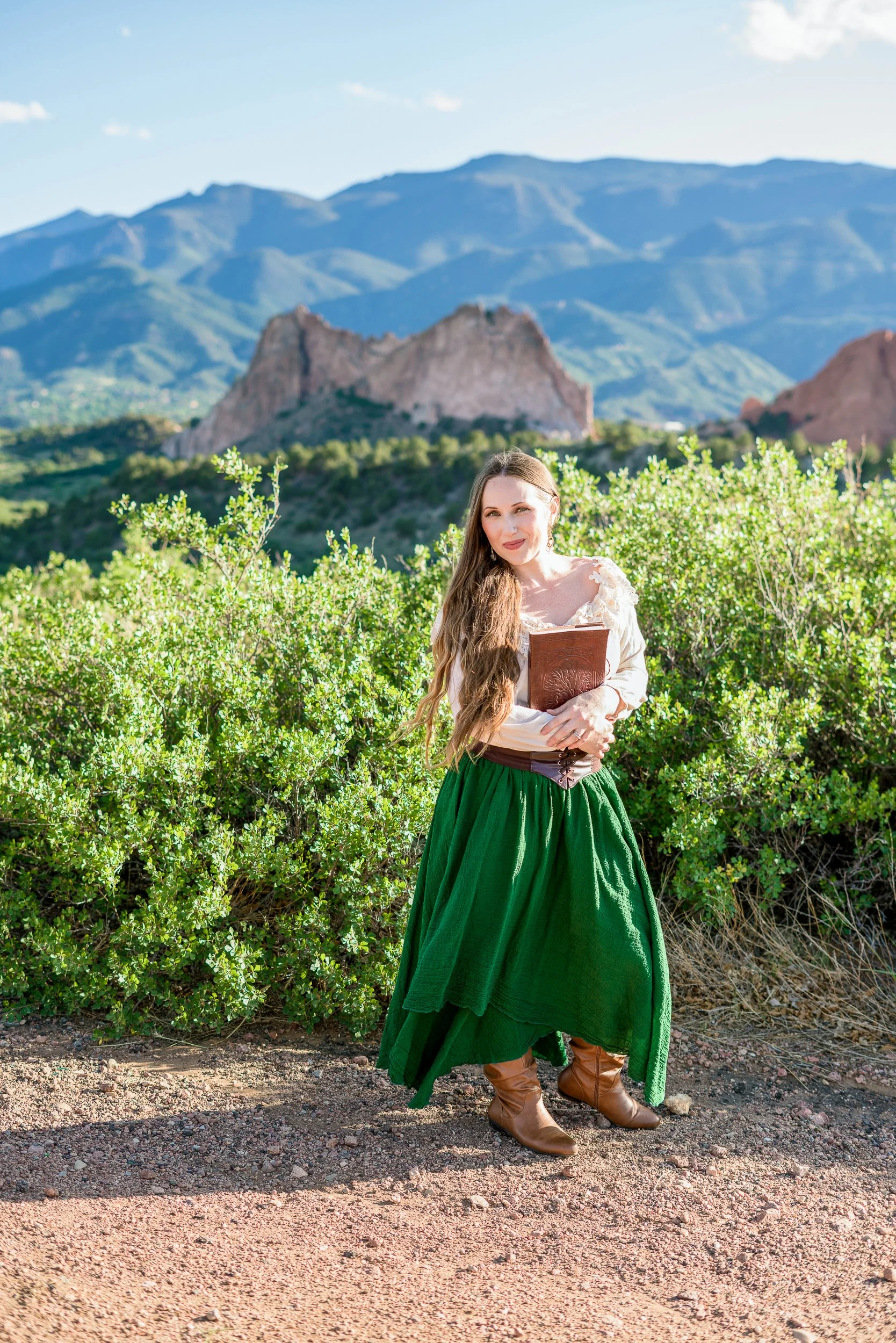Woman in a green skirt, brown boots, and a cream blouse holding a book outdoors in Colorado Springs, with mountains and a blue sky in the background during an author branding session.
