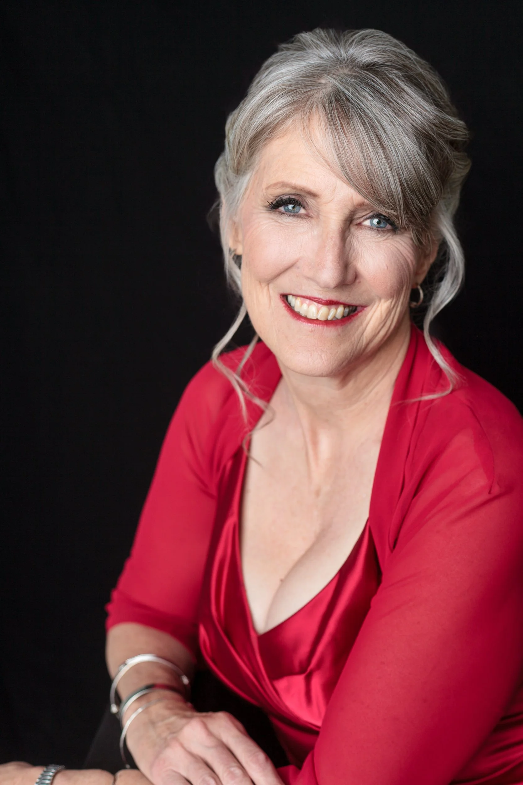 A smiling mature woman with short gray hair, blue eyes, wearing a red dress, silver jewelry, against a black background.