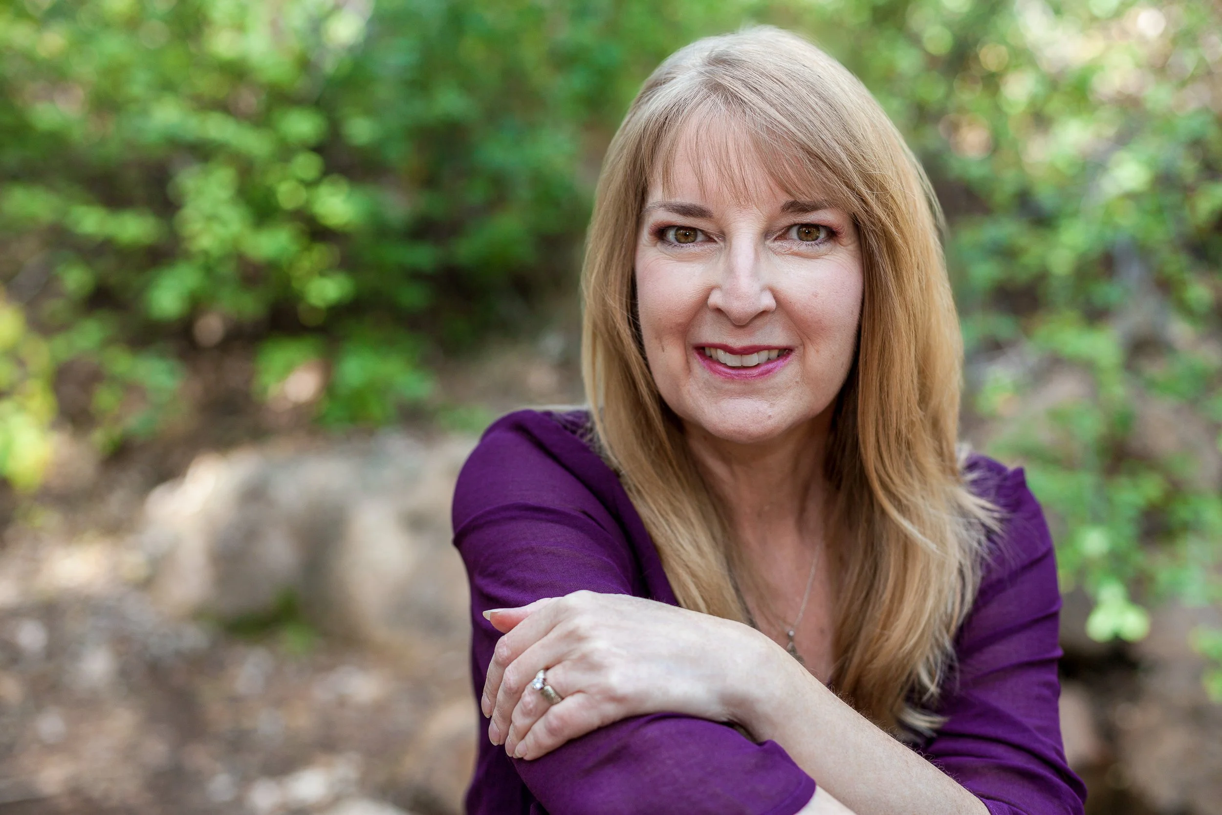 Smiling woman in a purple top with blonde hair, photographed outdoors in Colorado Springs with a soft green background.