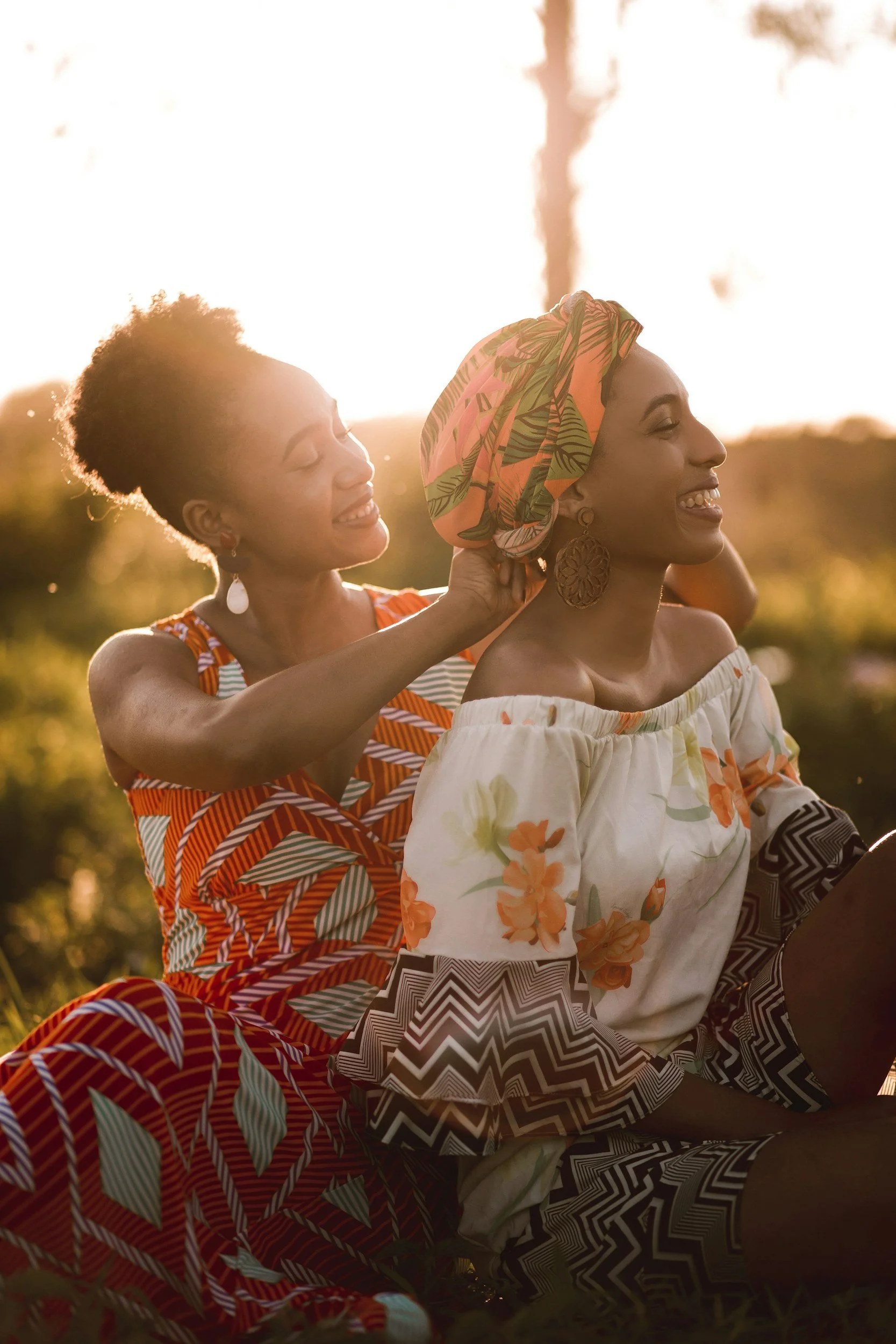 Two women enjoying a sunny afternoon outdoors; one adjusts a colorful headscarf on the other, both smiling and wearing vibrant, patterned dresses.