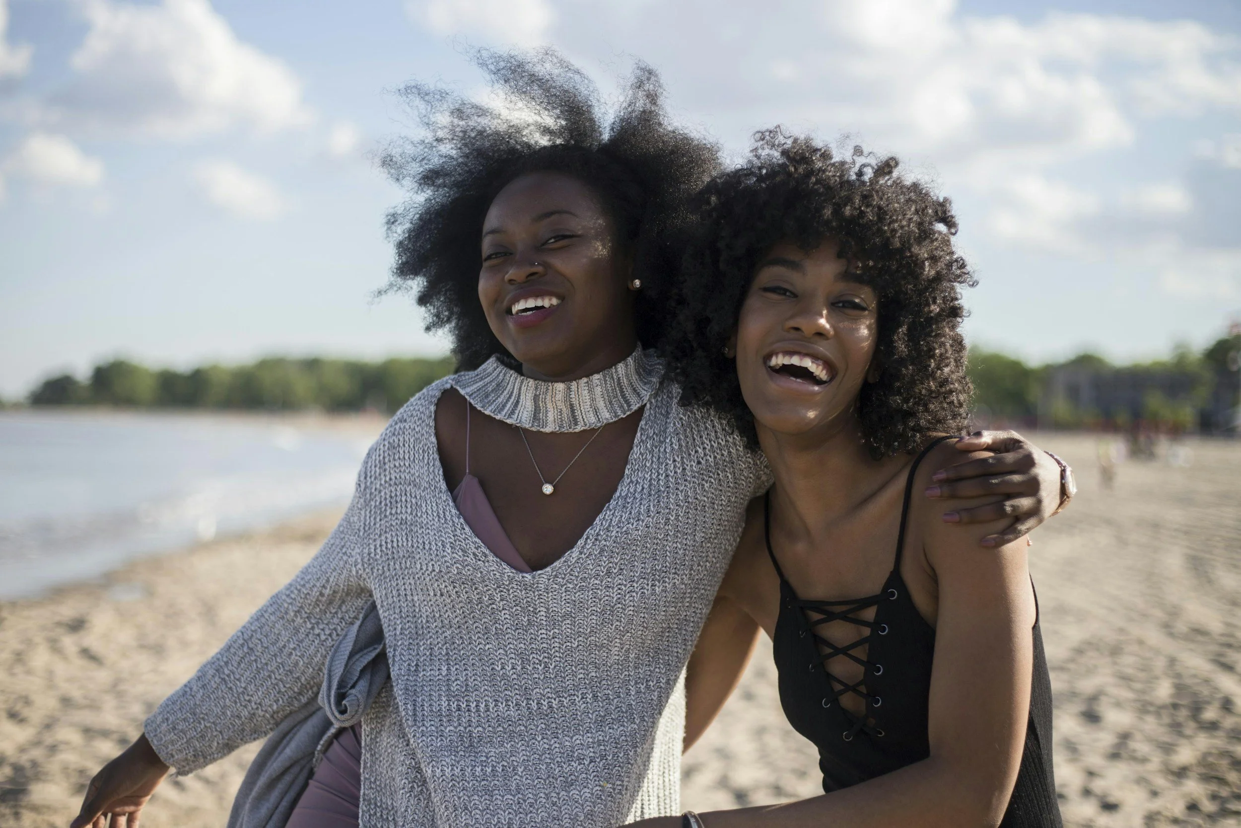 Two women smiling and embracing on a beach, with the ocean and a partly cloudy sky in the background.