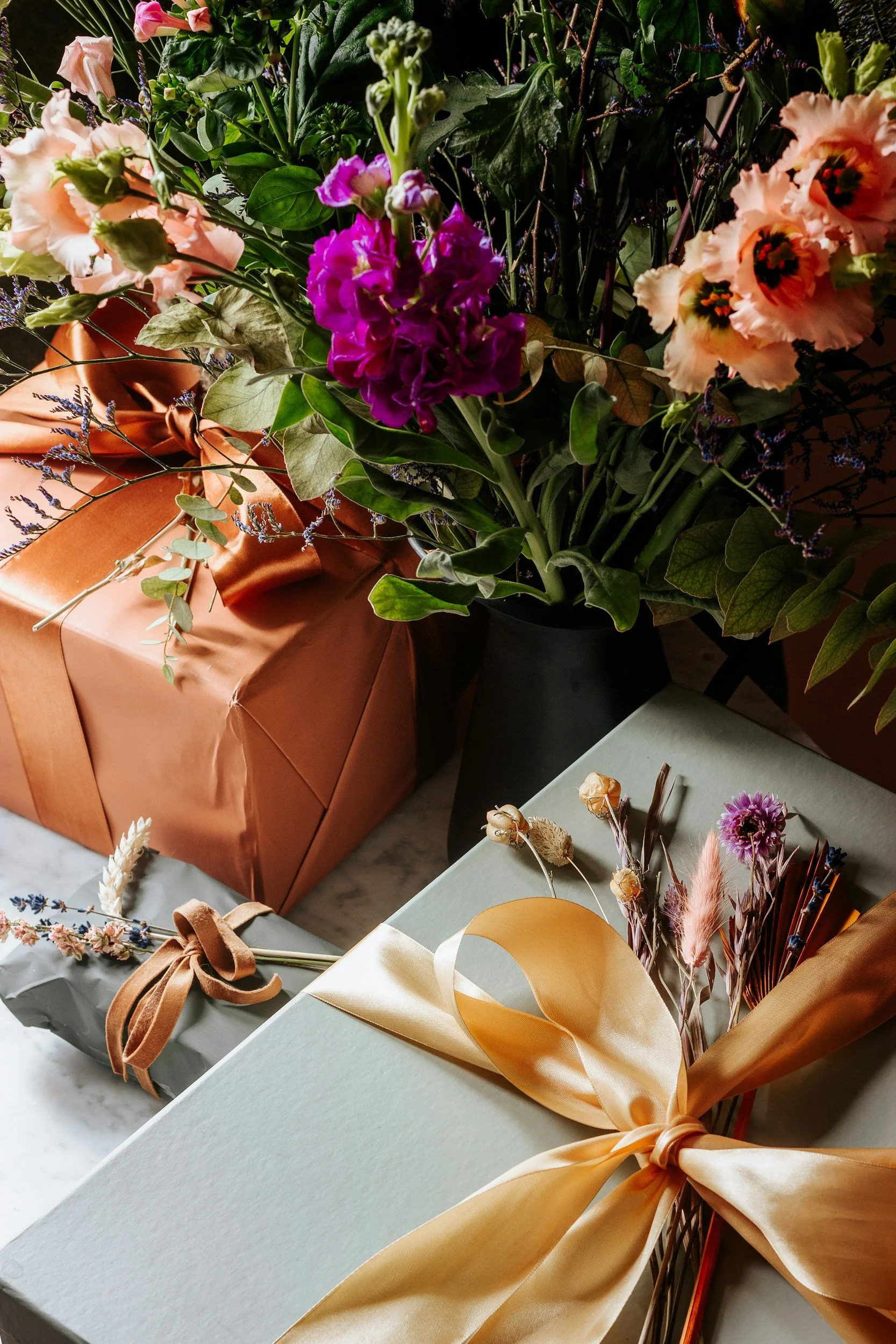 An arrangement of wrapped gift boxes and a bouquet of colorful flowers, including pink, purple, and cream-colored blossoms, all placed on a marble surface.