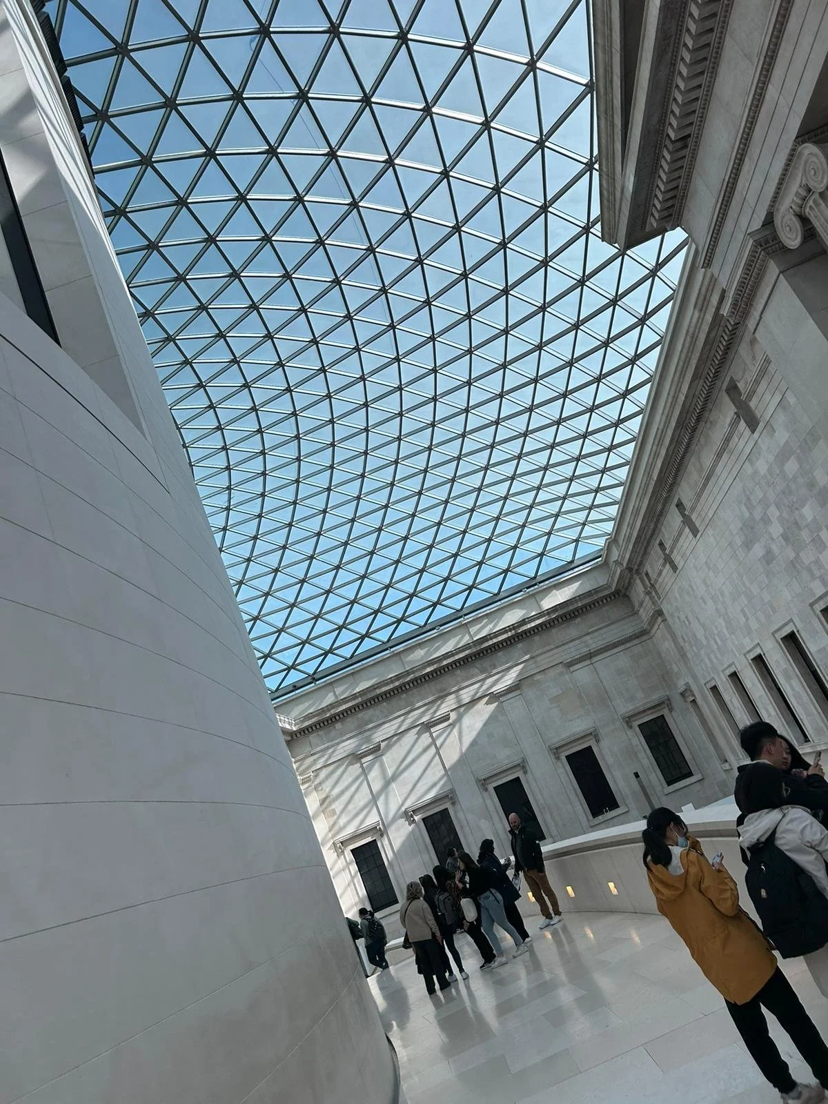 Inside a museum with a glass dome ceiling, people are walking and standing along a curved wall, some looking at their phones.