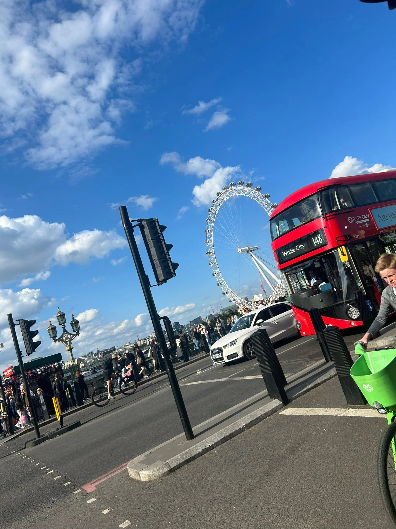 A busy street scene in London featuring a red double-decker bus, a white car, pedestrians, a Ferris wheel, and a clear blue sky with scattered clouds.