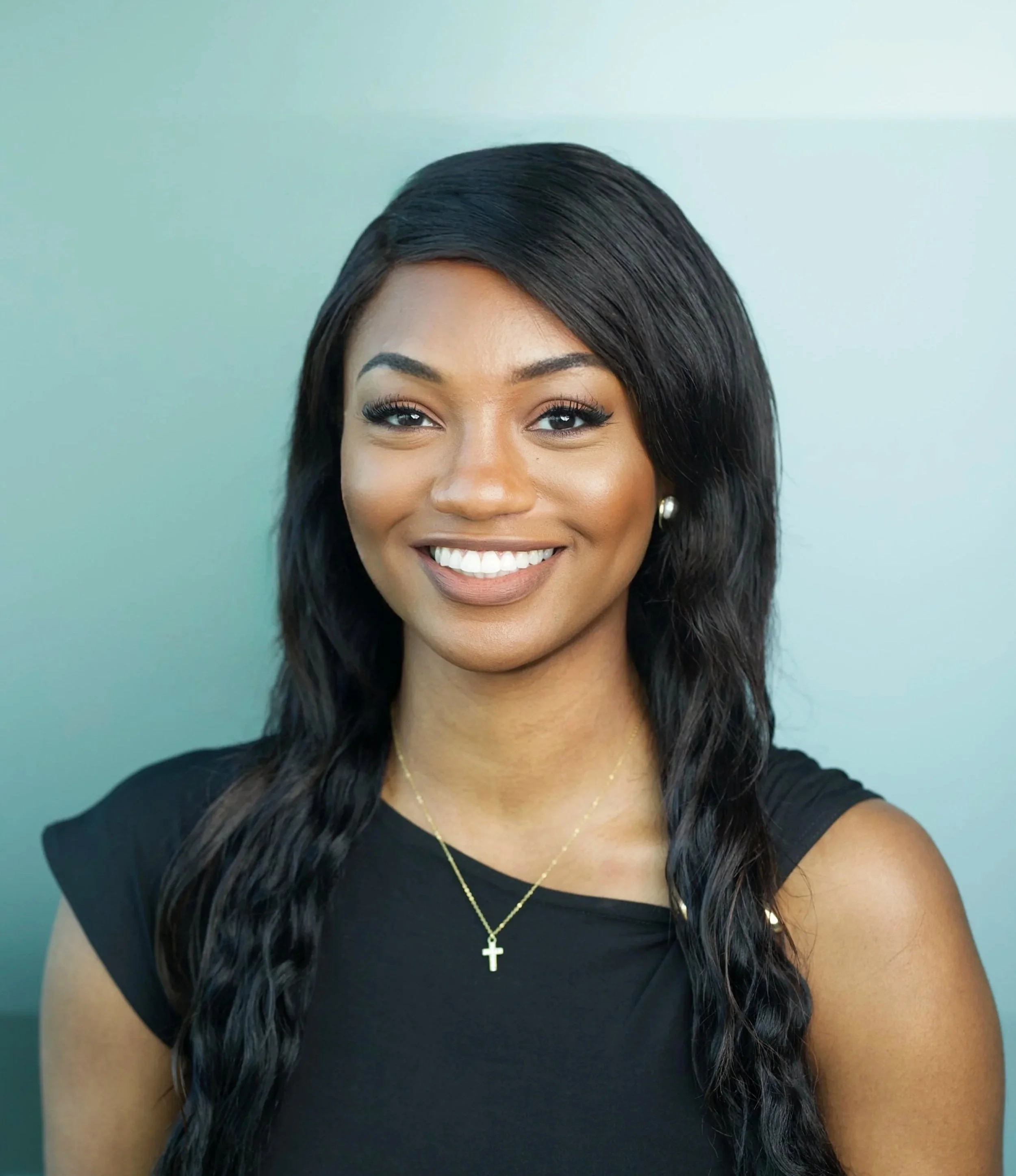Portrait of a young African American woman smiling, wearing a black top and gold jewelry, against a plain background.