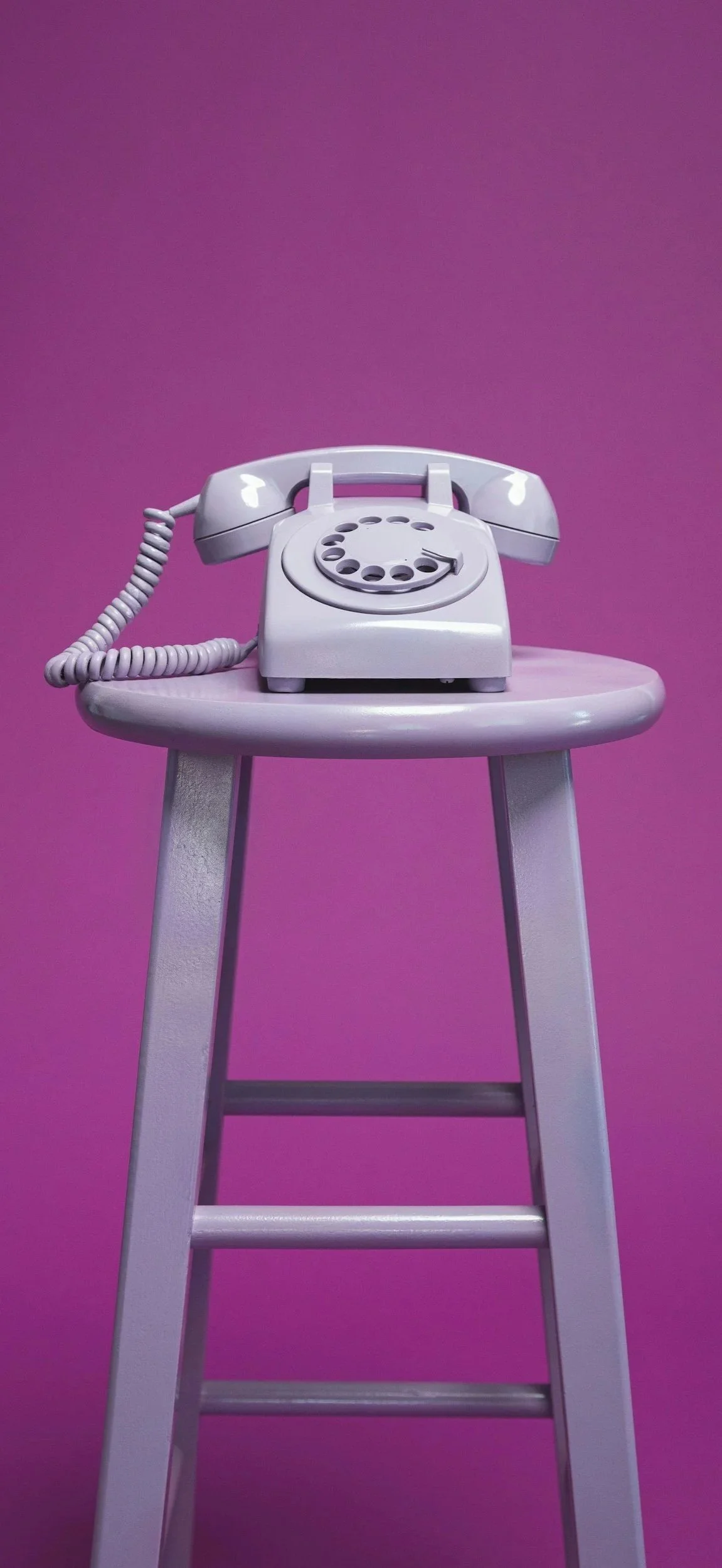 A white rotary telephone placed on a small white stool against a purple background.