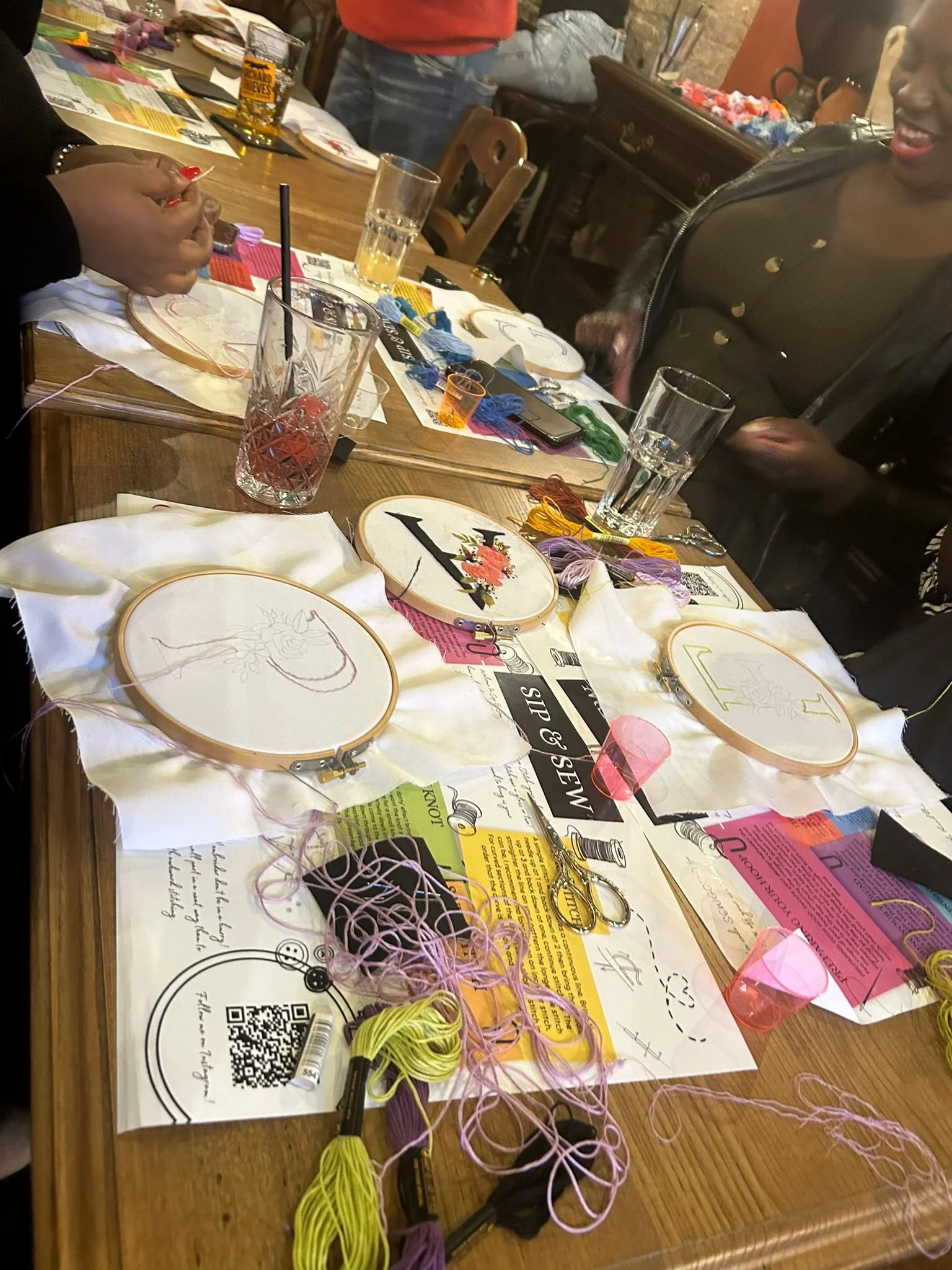 A table with embroidery hoops, colored threads, scissors, and crafting supplies, with people gathered around preparing for embroidery craft activity.