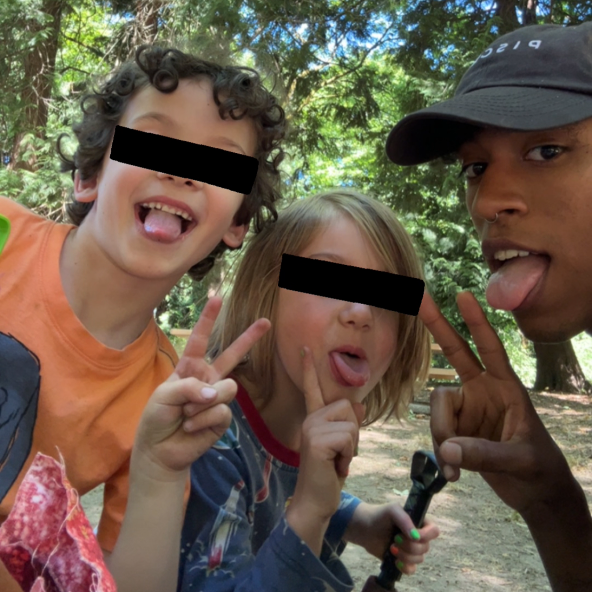 Three kids outdoors making silly faces, with two sticking out their tongues and one showing a peace sign, in a wooded area.