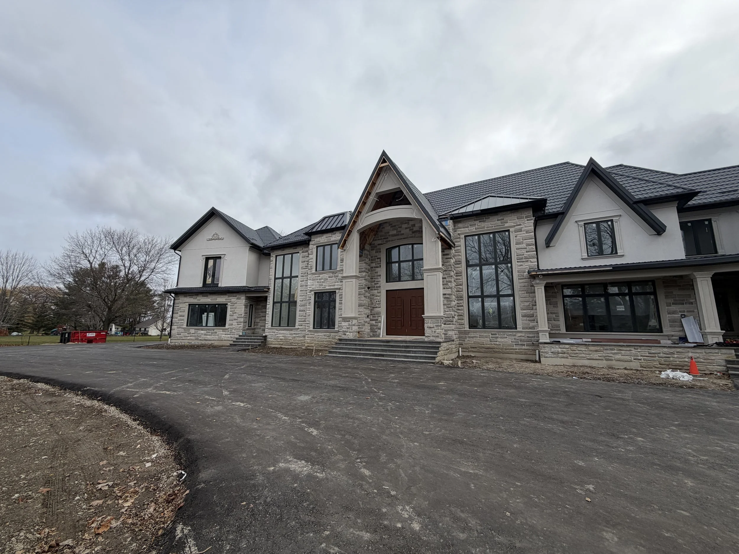 A large, modern house under construction with a paved driveway and a cloudy sky overhead.