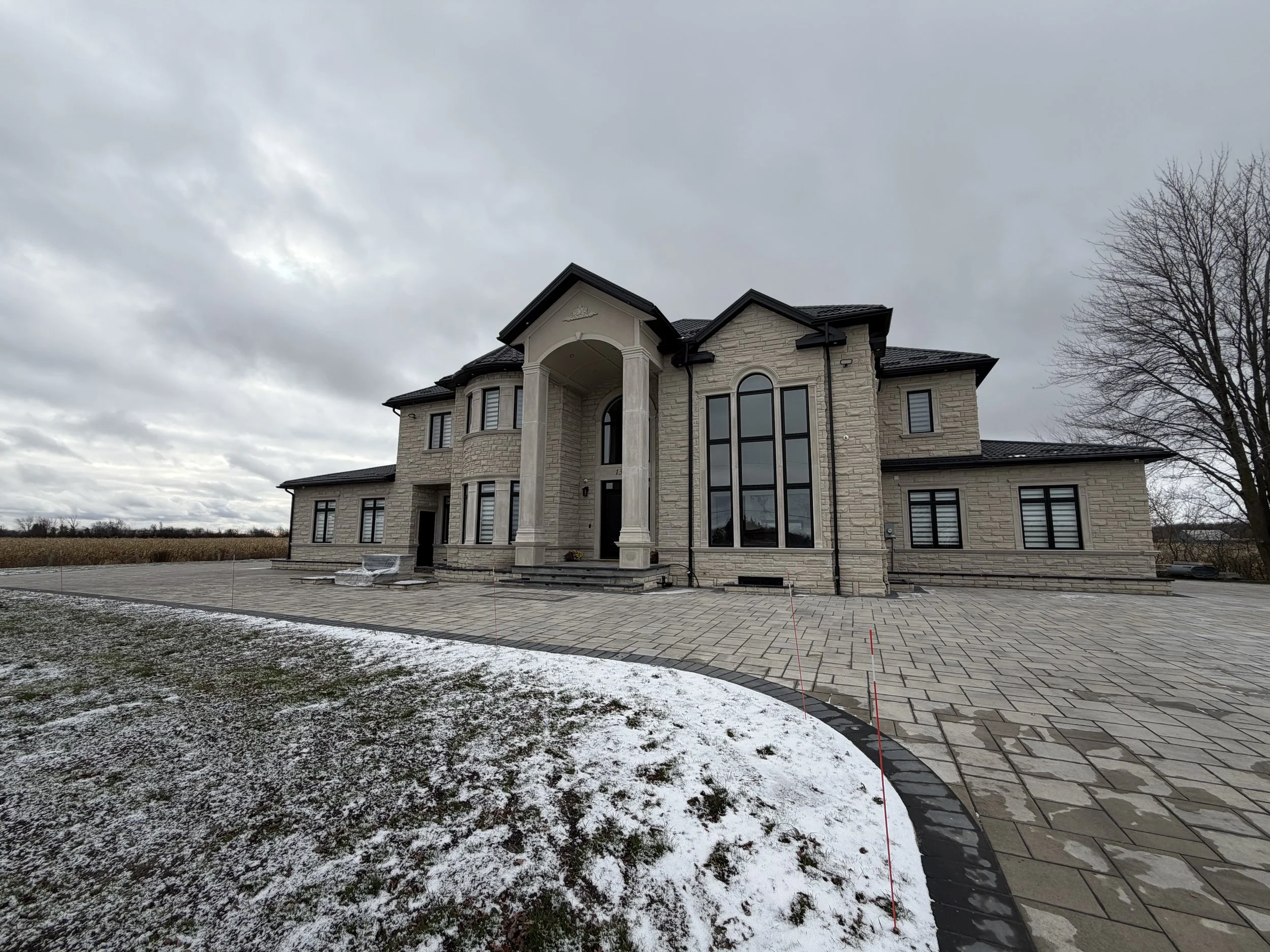 Large beige stone mansion with black roof and multiple windows, a spacious stone driveway, snowy patches on the ground, on a cloudy day with overcast sky, and leafless tree on the right.