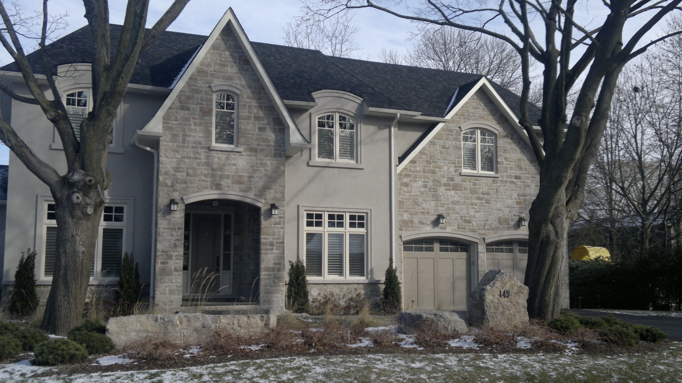Front view of a two-story house with a stone facade, white trim, and a dark shingle roof, with two large trees in the yard and a small patch of snow on the ground.