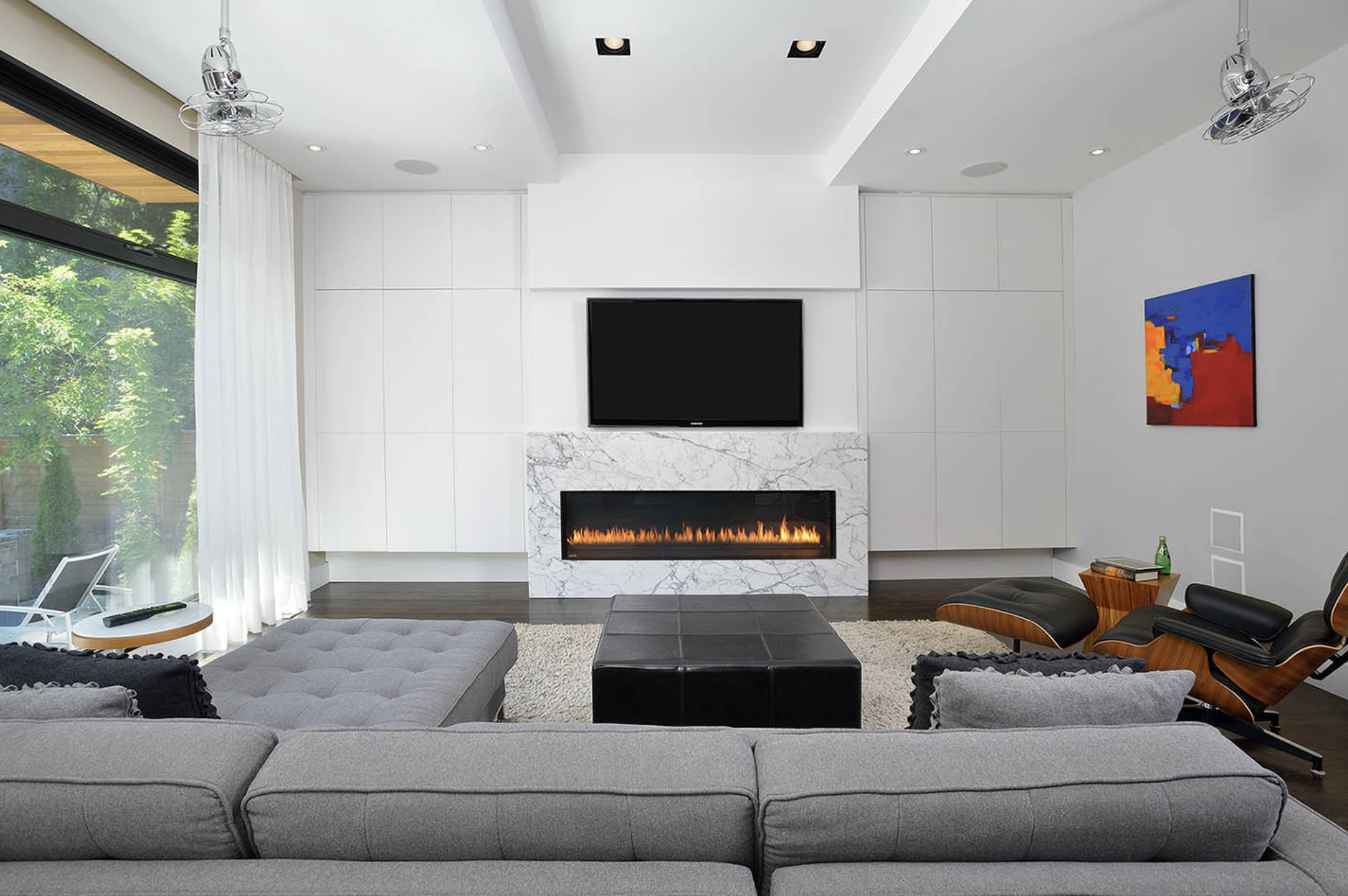 Modern living room with gray sofa, black ottoman, white marble fireplace, mounted TV, colorful abstract artwork, side table with a bottle, large window with white curtains, ceiling fans, and a view of greenery outside.